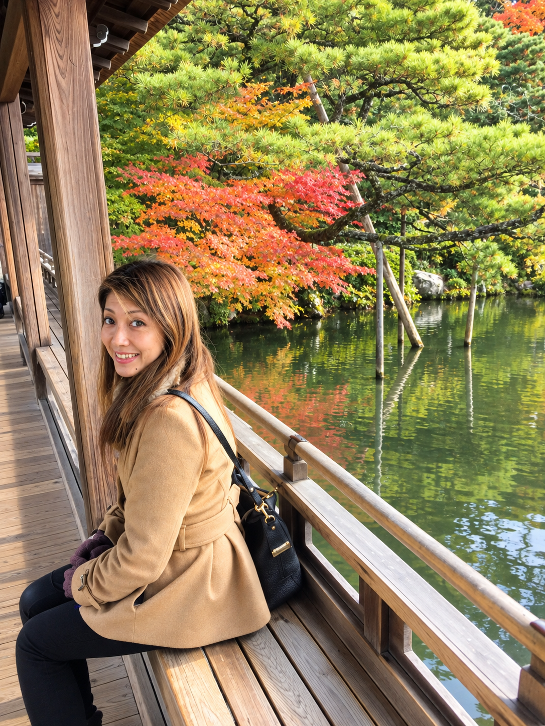 A woman sitting on a wooden bench on a bridge or balcony overlooking a pond with colorful autumn trees in the background.