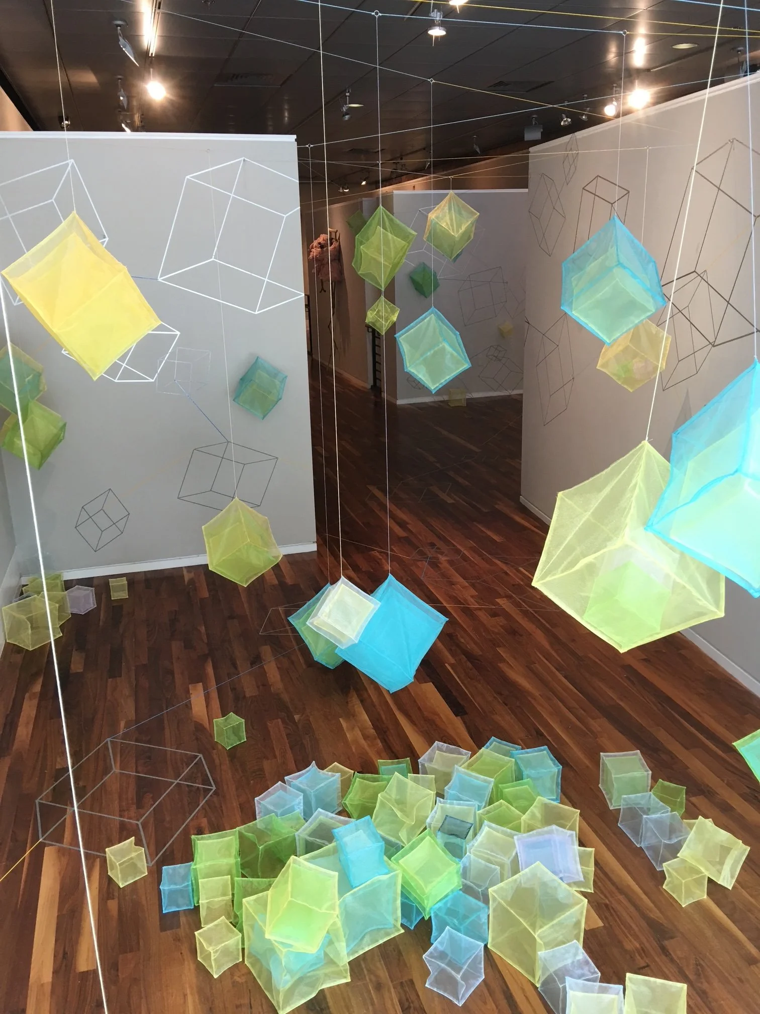 An art installation featuring colorful, translucent geometric shapes and wireframe cube outlines hanging from the ceiling in an indoor space with wooden flooring.