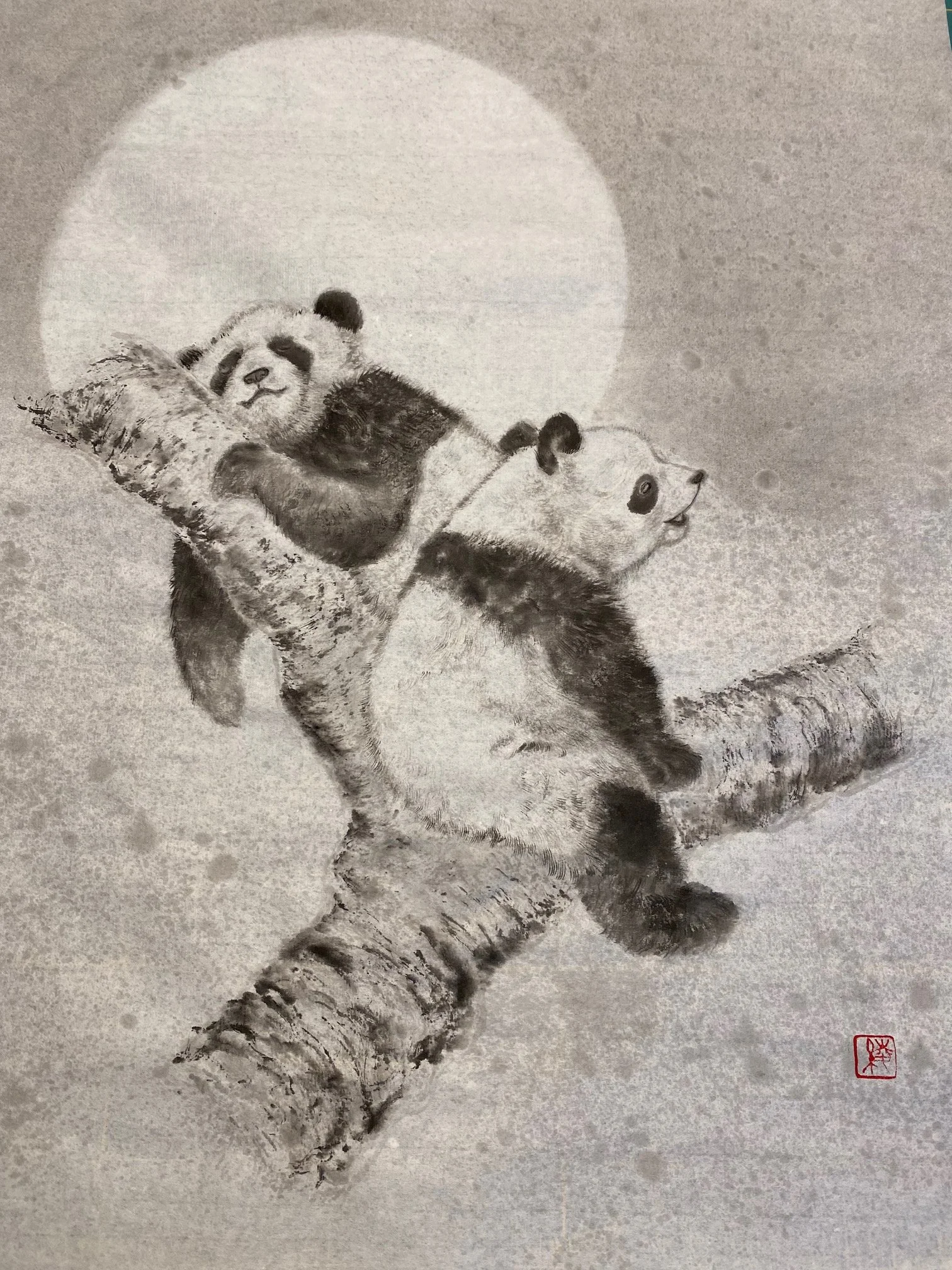 A traditional black and white ink painting of two pandas resting on a tree branch with a full moon in the background, signed with a red artist's seal.