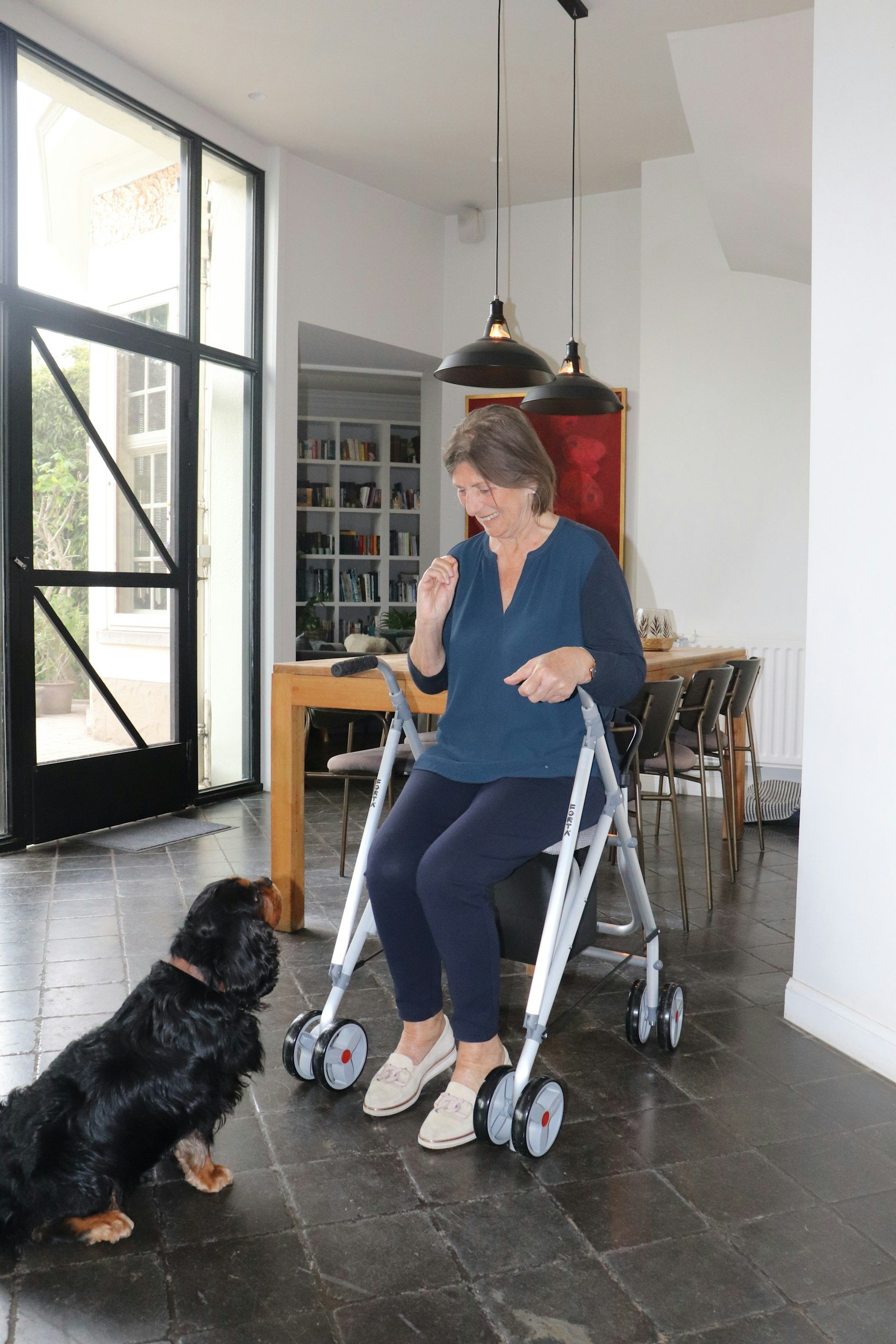 An elderly woman with short gray hair, glasses, wearing a blue top, dark pants, and slippers, sitting on a walker in a modern kitchen or dining area. She is smiling and looking at a black and tan dog, a puppy, sitting on the floor and looking up at her. The room has large windows, dark tile flooring, a wooden dining table, chairs, and a bookshelf in the background.