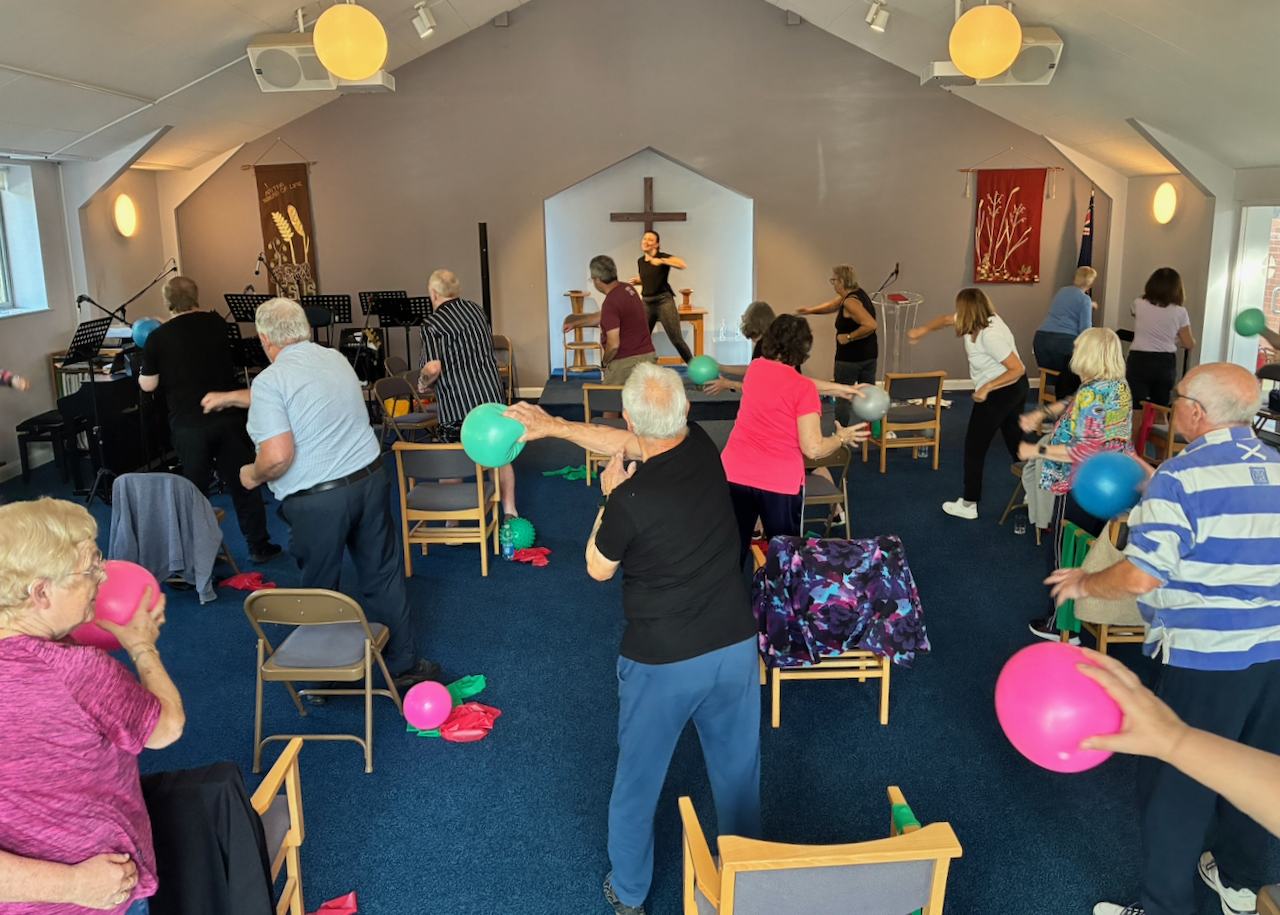 Group of elderly people participating in an exercise class in a church with a cross on the wall. They are holding colorful exercise balls and following a leader in front of the congregation.