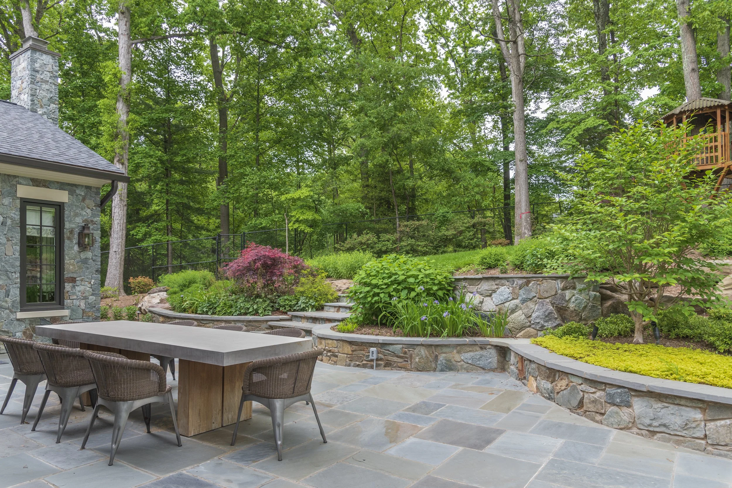 A well-manicured backyard patio with a large stone table and chairs, stone retaining walls, lush greenery including shrubs and trees, and a wooded area in the background.