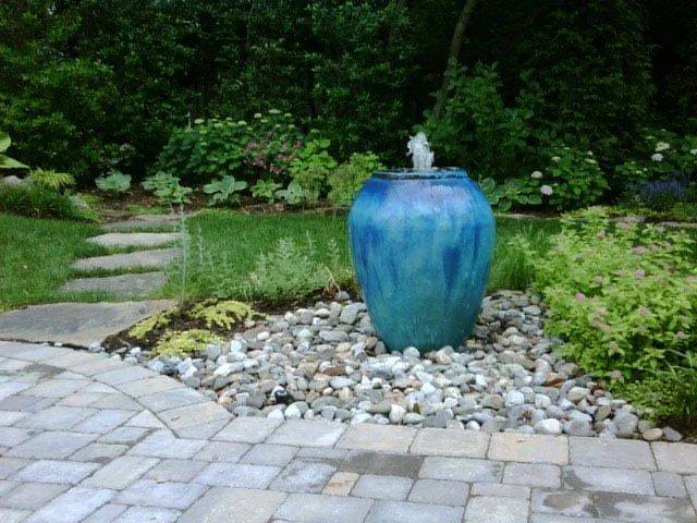 Bright, colorful water feature with a ceramic urn. Texture from lush plantings, smooth river stones, and cobblestone style concrete pavers in McLean, VA