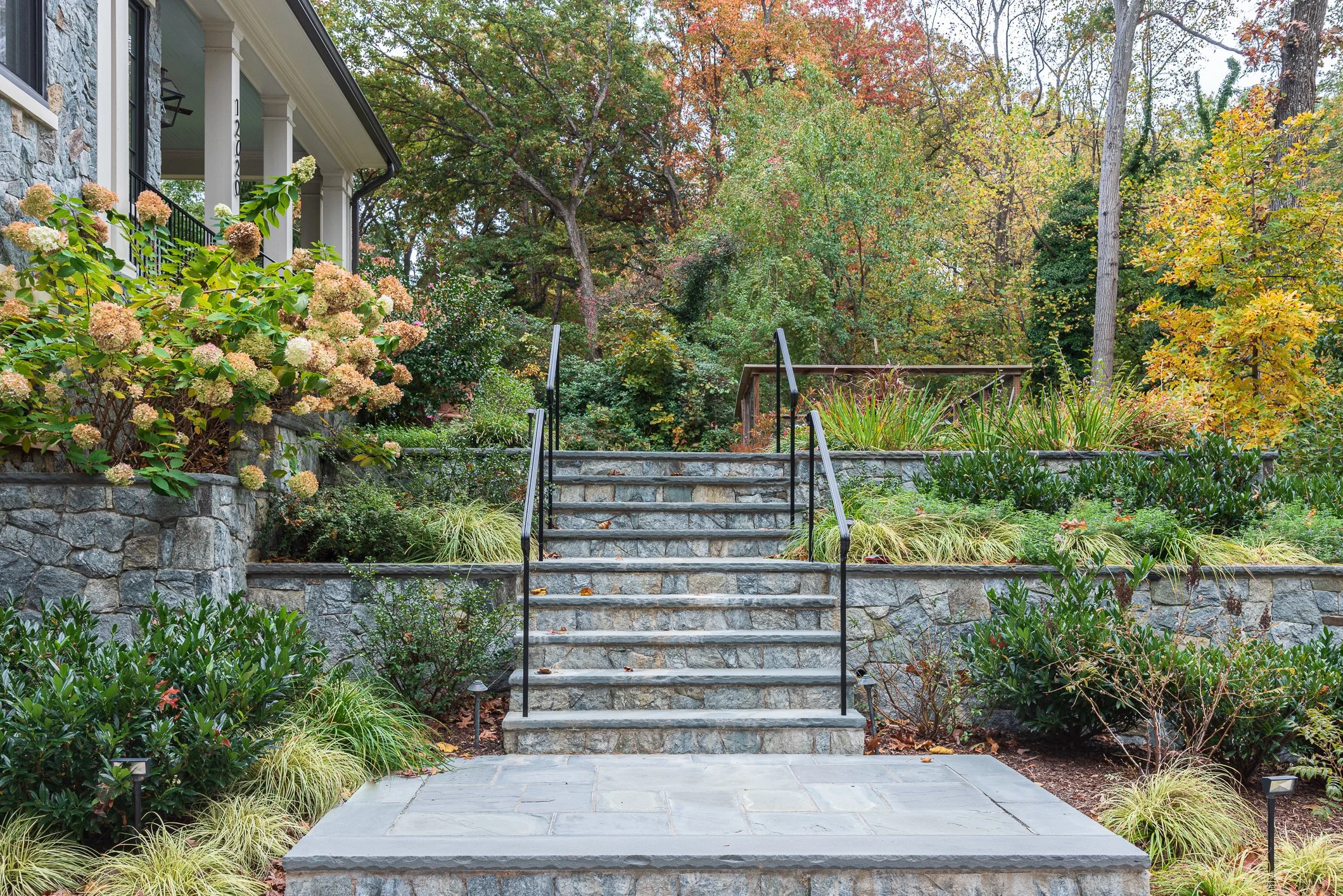 Stone steps and retaining walls for a steep sloped front yard. Opening and welcoming design for a front entry. Colorful plantings compliment the mature native trees fall foliage