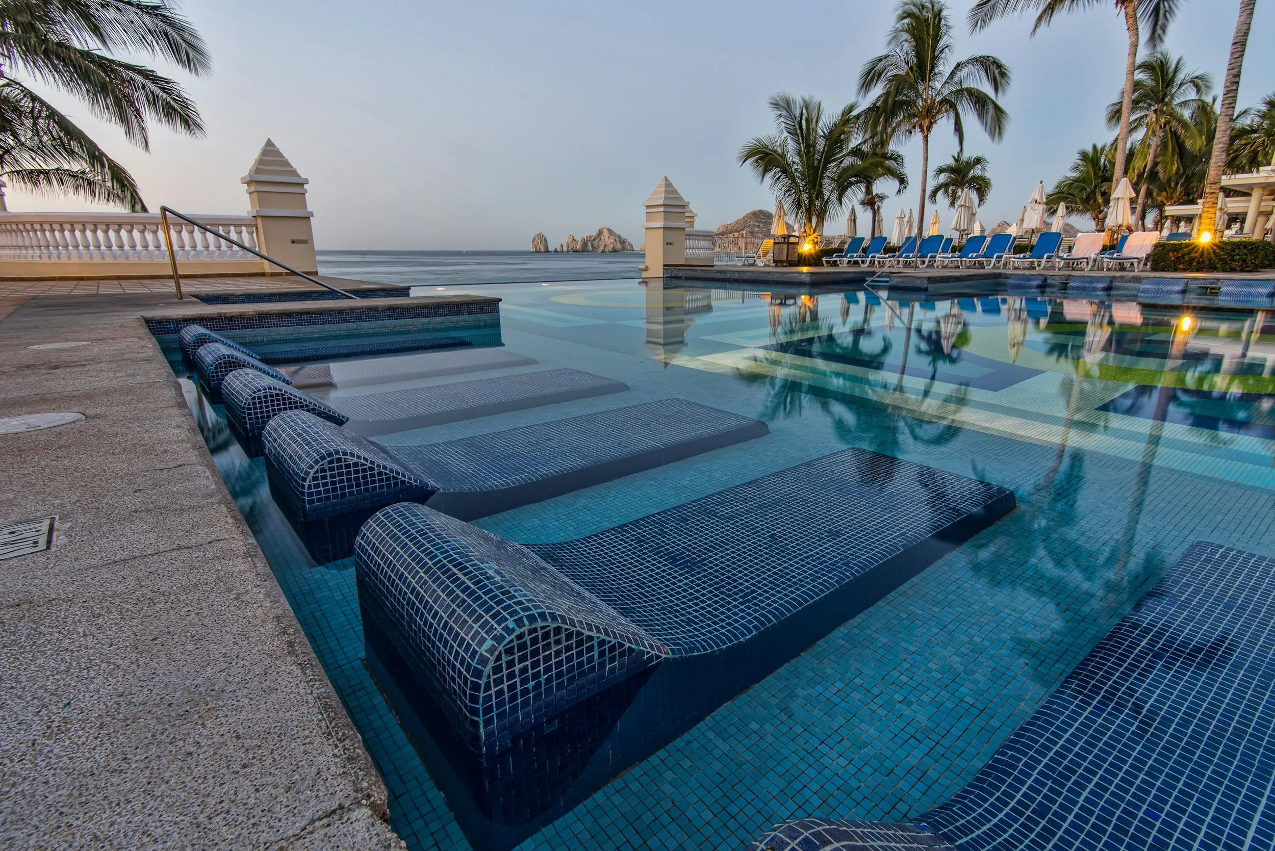 A luxurious poolside area with an infinity pool overlooking the ocean, surrounded by palm trees, lounge chairs, white umbrellas, and small islands in the distance at sunset.