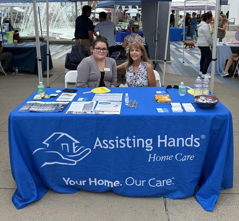 Two women sitting at a table covered with a blue tablecloth that says "Assisting Hands Home Care" and "Your Home. Our Care." inside a tent at an outdoor event.