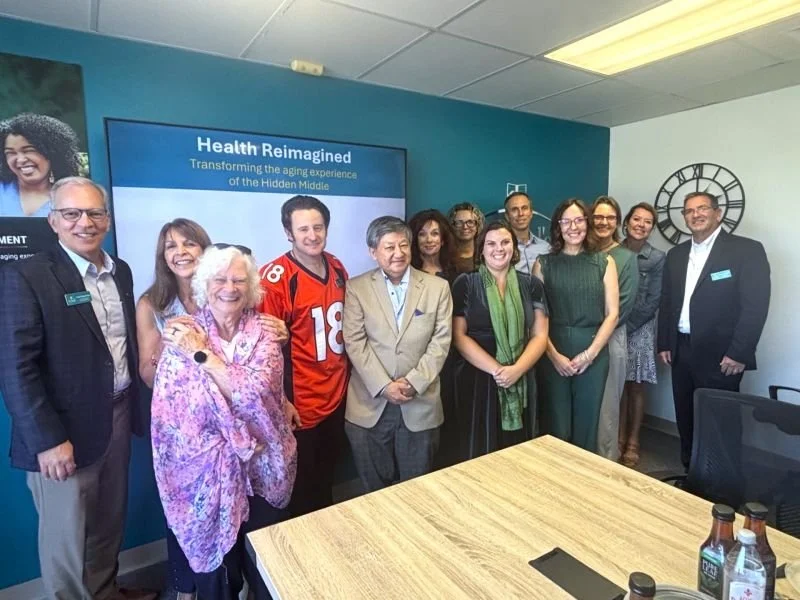 A group of people posing in a conference room with a sign that reads 'Health Reimagined: Transforming the aging experience of the Hidden Middle.'