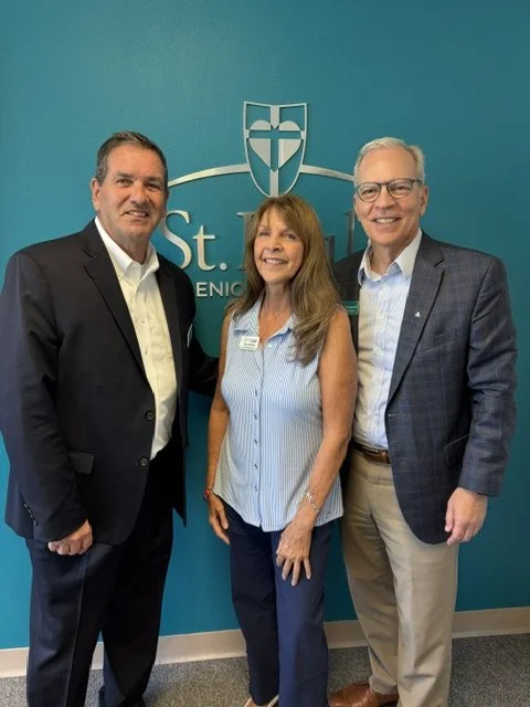 Three adults standing in front of a teal wall with a St. Elizabeth logo, smiling at the camera.