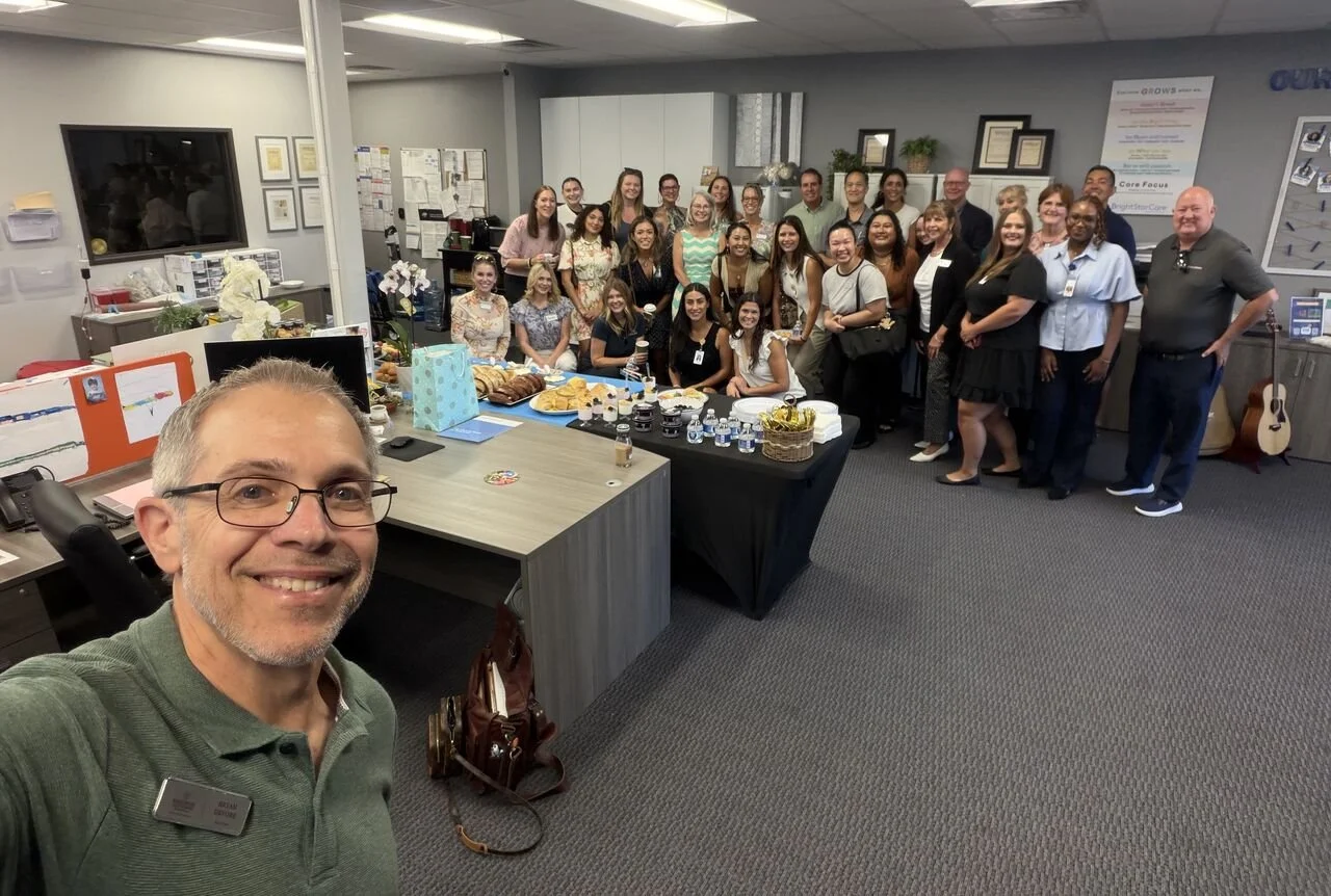 A group of people gathered in an office, posing for a group photo during a celebration or event. The office has a table with snacks and drinks, and a man taking a selfie at the front in a green shirt and glasses.