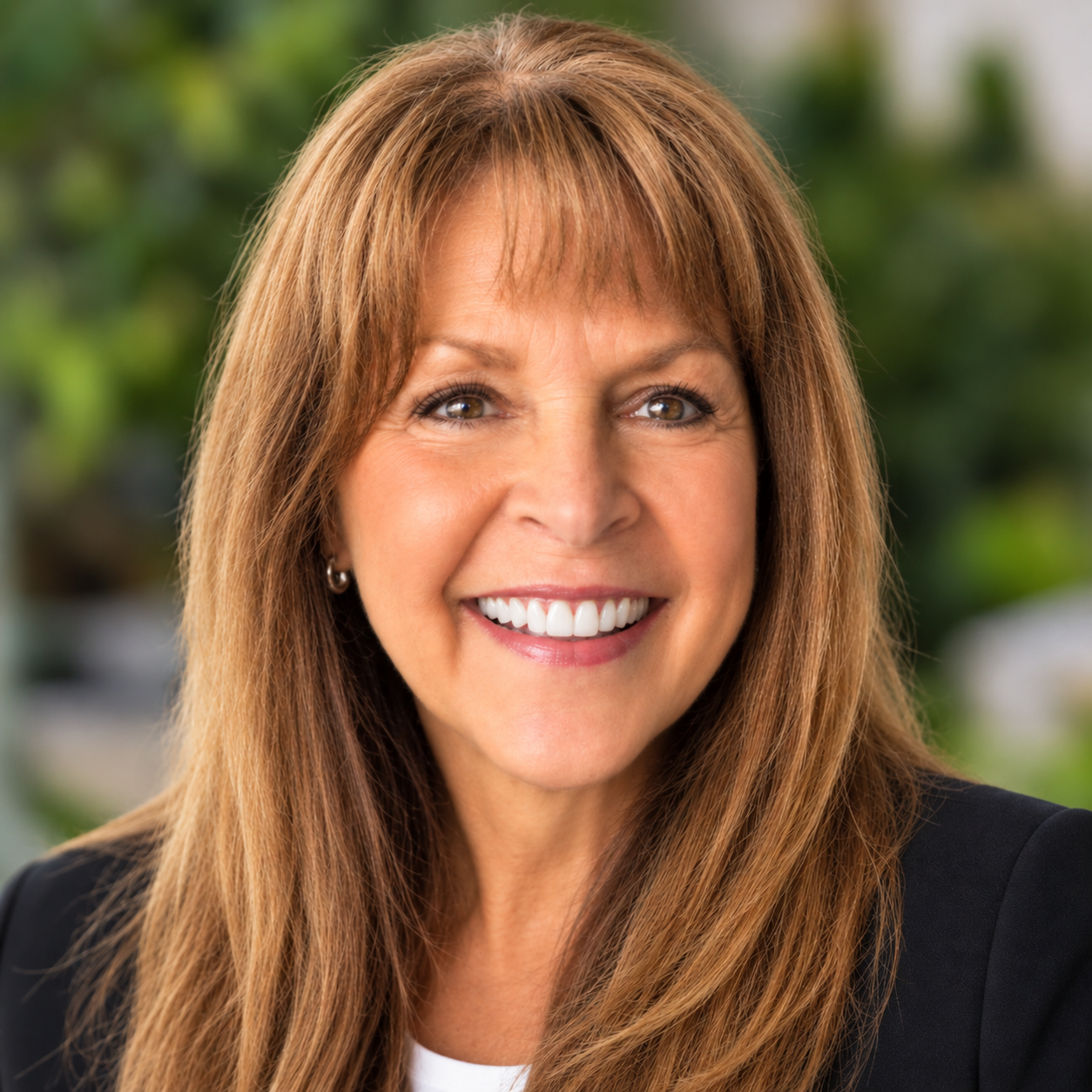 Close-up of a smiling woman with long reddish-brown hair, wearing a black blazer outdoors with green foliage in the background.
