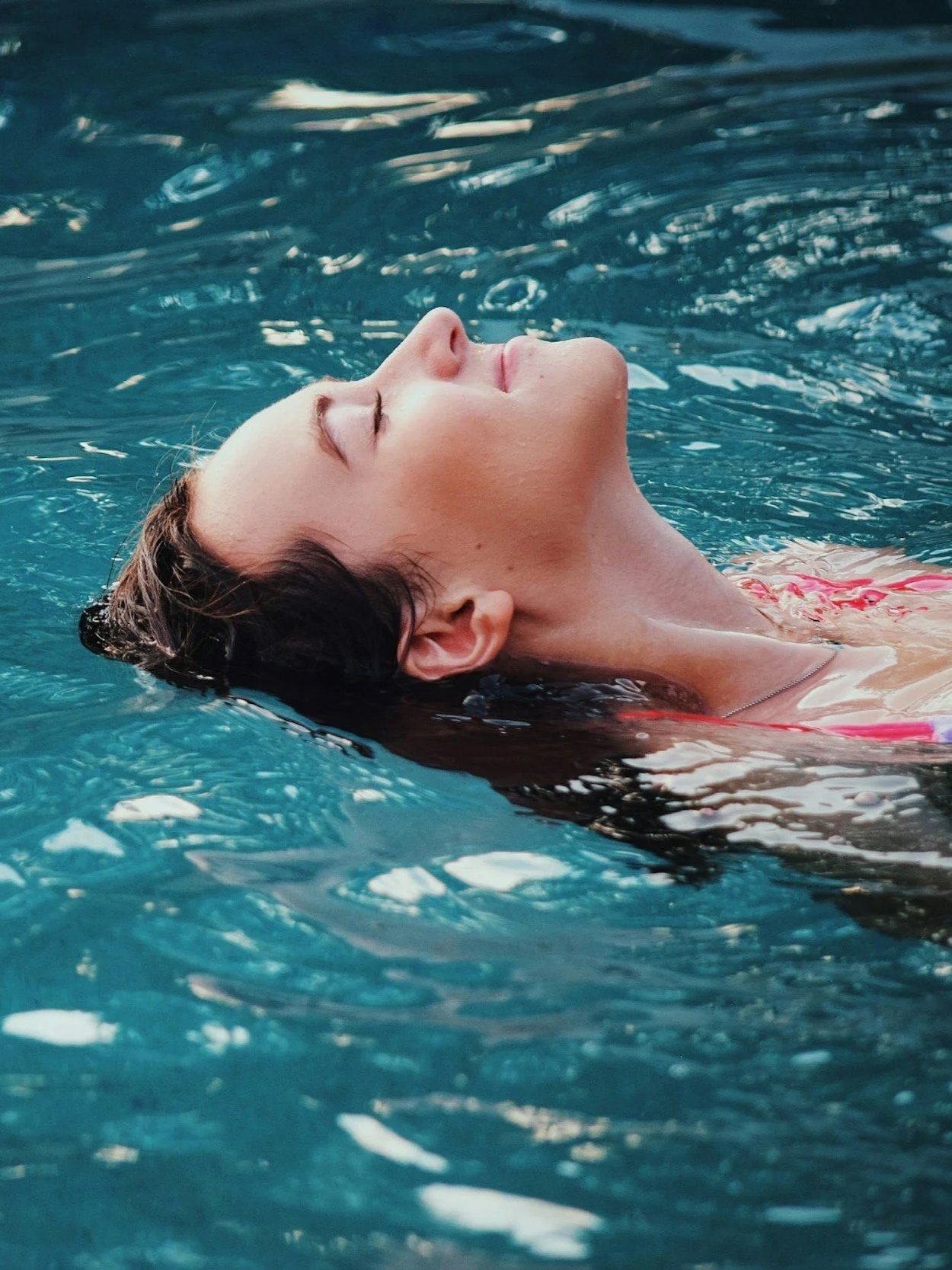 Eine Frau entspannt im Wasser mit geschlossenen Augen und geneigtem Kopf, im Pool schwimmend, trägt ein rotes Badeoberteil.