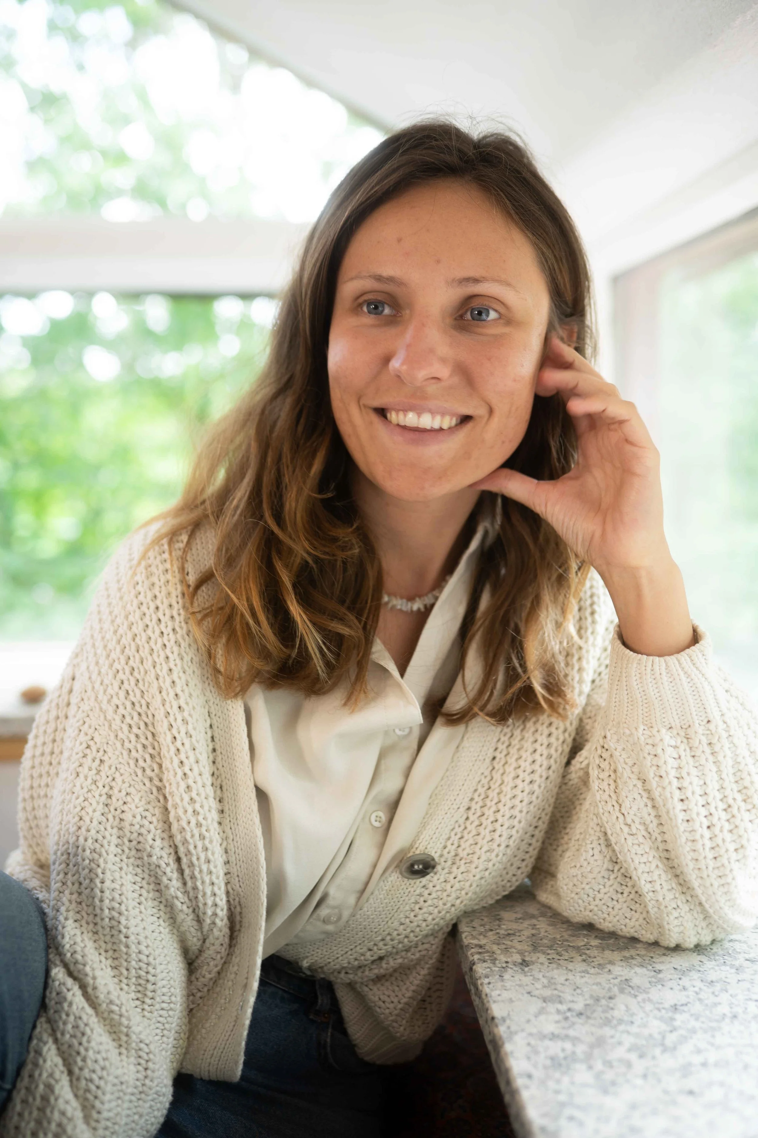 Junge Frau mit braunen, gewellten Haaren sitzt an einer Steintheke, trägt einen weißen Hemd und einen beigefarbenen, gestrickten Pullover, lächelt und berührt ihr Ohr, im Hintergrund ist viel natürliches Licht und grüne Bäume sichtbar.