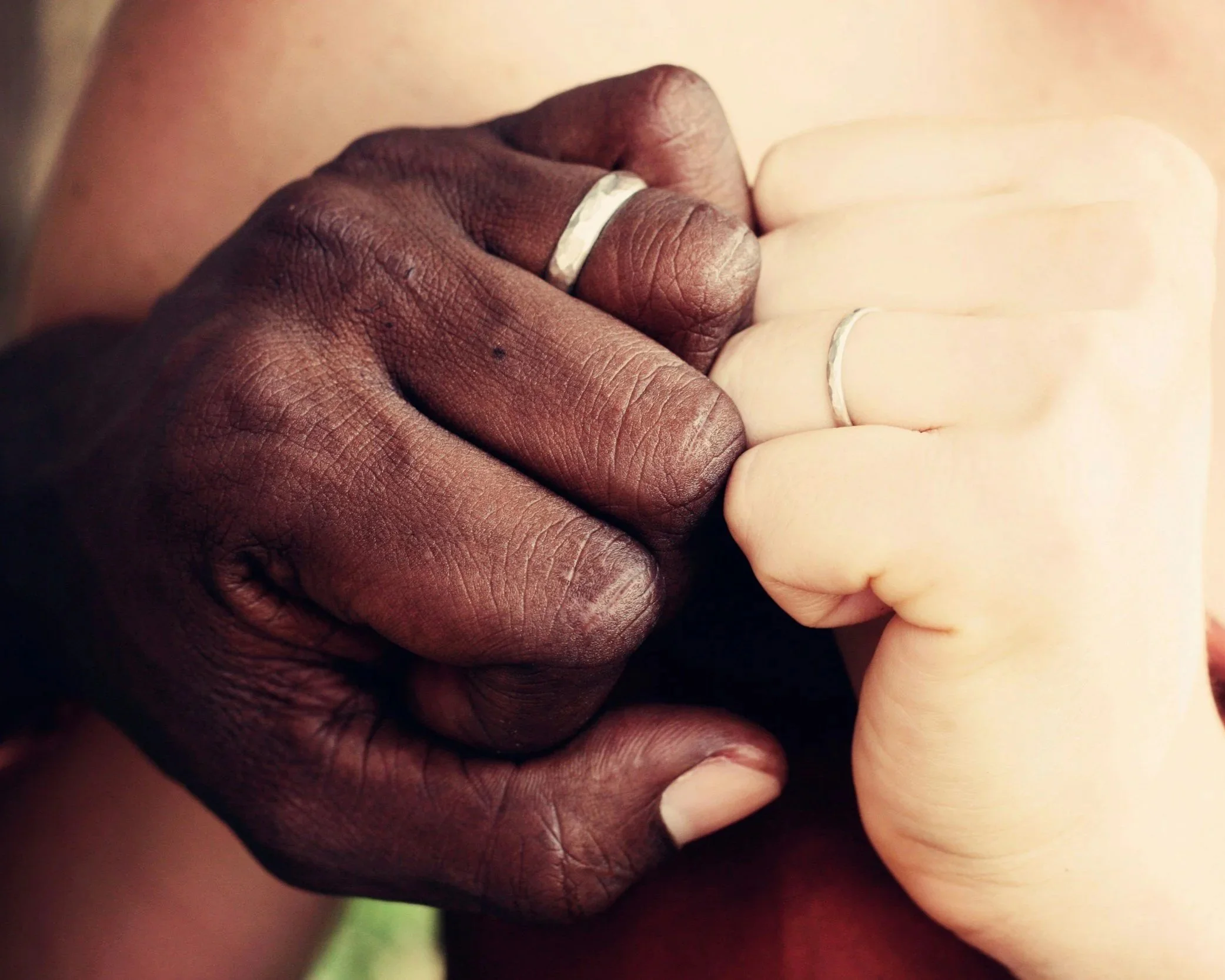 Close-up of two hands, one dark-skinned and one light-skinned, clasped together with rings on their fingers.