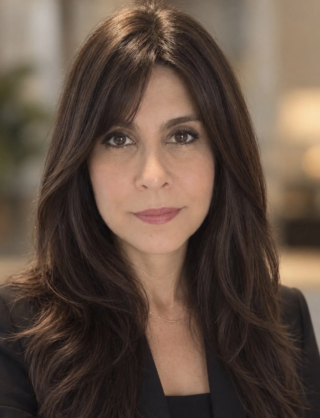 A woman with long brown hair and brown eyes, wearing a black blazer and a delicate necklace, smiling softly at the camera in an indoor setting.