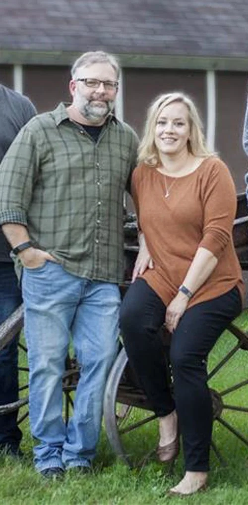 A man with glasses wearing a green plaid shirt and jeans, standing next to a woman with blonde hair in a rust-colored top and black pants, sitting on a wooden wagon or cart outdoors.