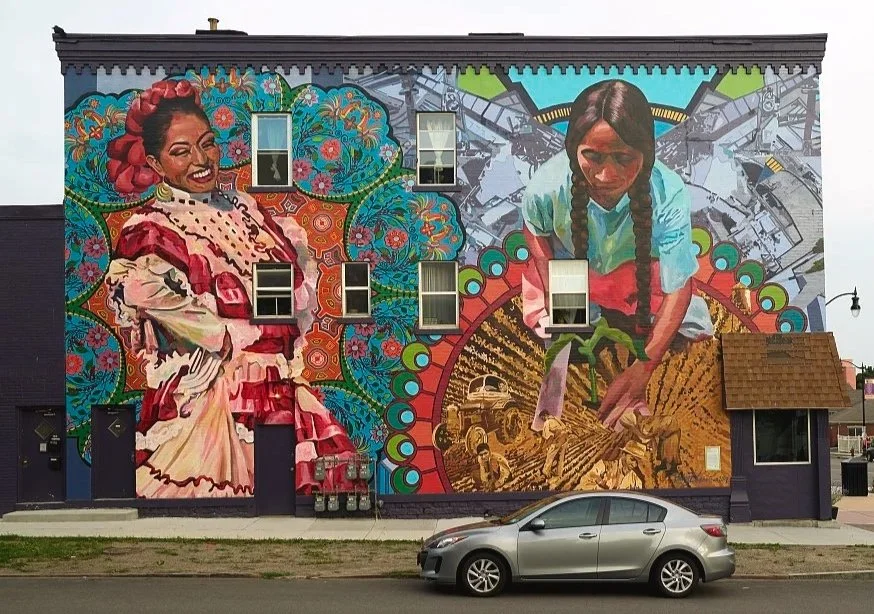 a mural of two figures, one woman wearing a white dress, another In a teal shirt. in front of the mural is a silver car.