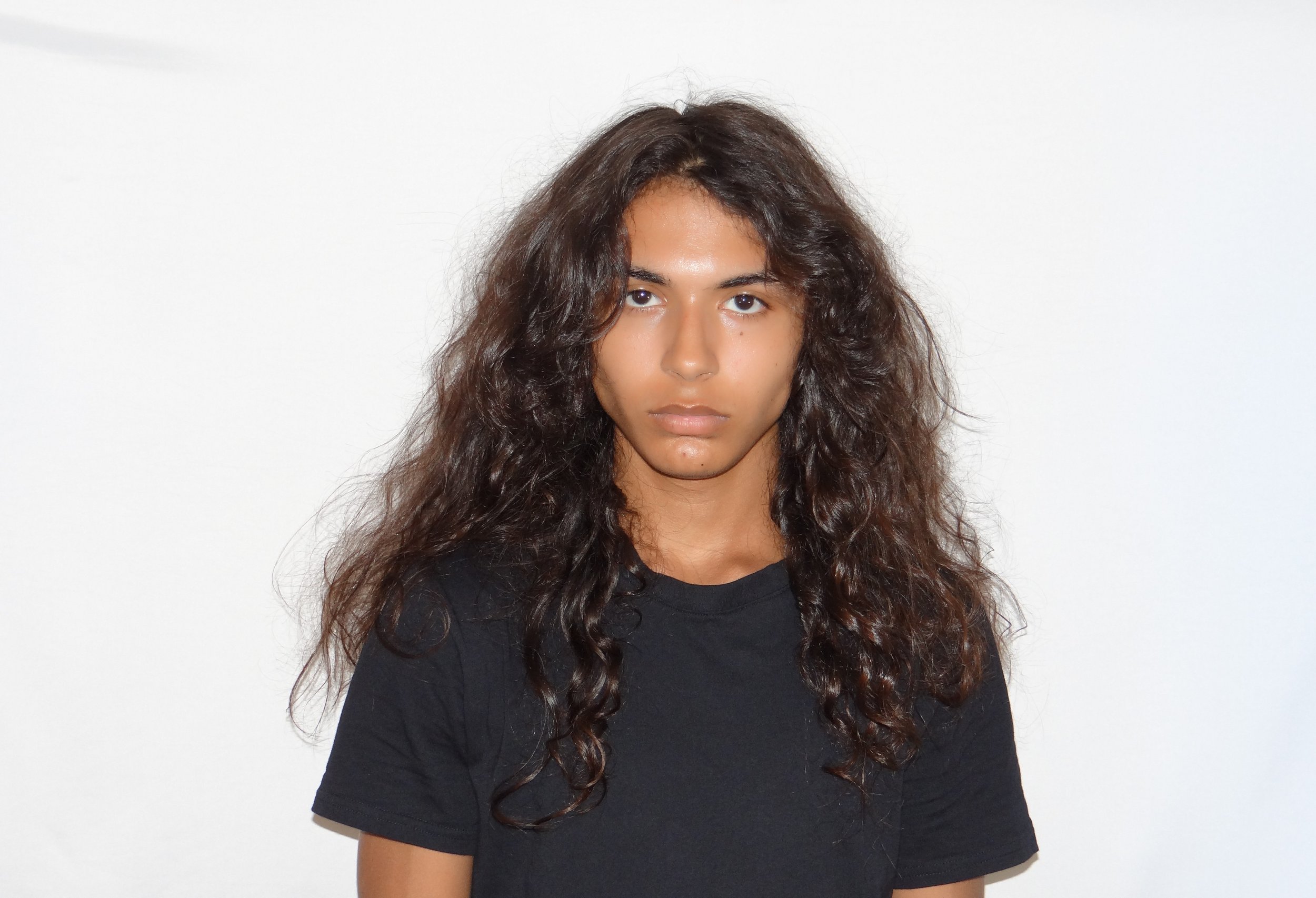 A young man with long, curly hair and tan skin looking directly at the camera against a plain white background, wearing a black t-shirt.