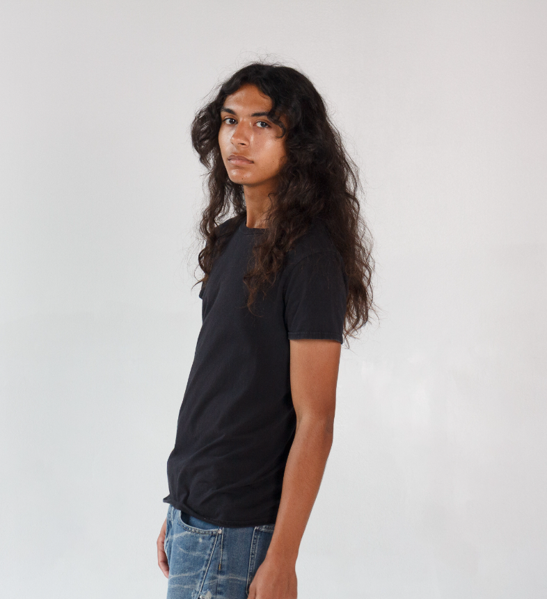 Portrait of a young man with long, curly hair wearing a black t-shirt and blue jeans, standing against a plain white background.