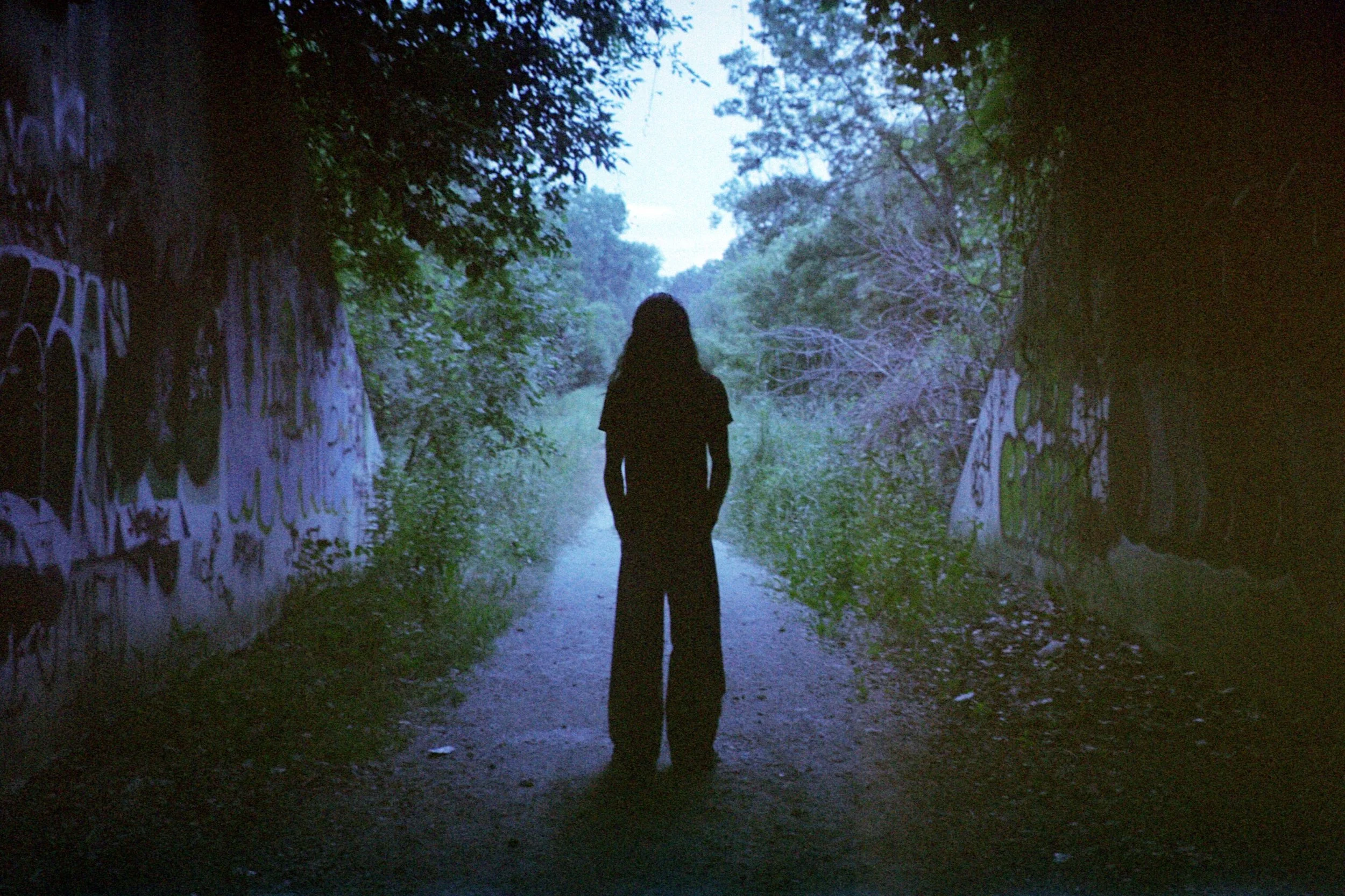 A person with long hair dressed in dark clothing walking through a tunnel with graffiti on the walls, leading to a bright, wooded outdoor area.