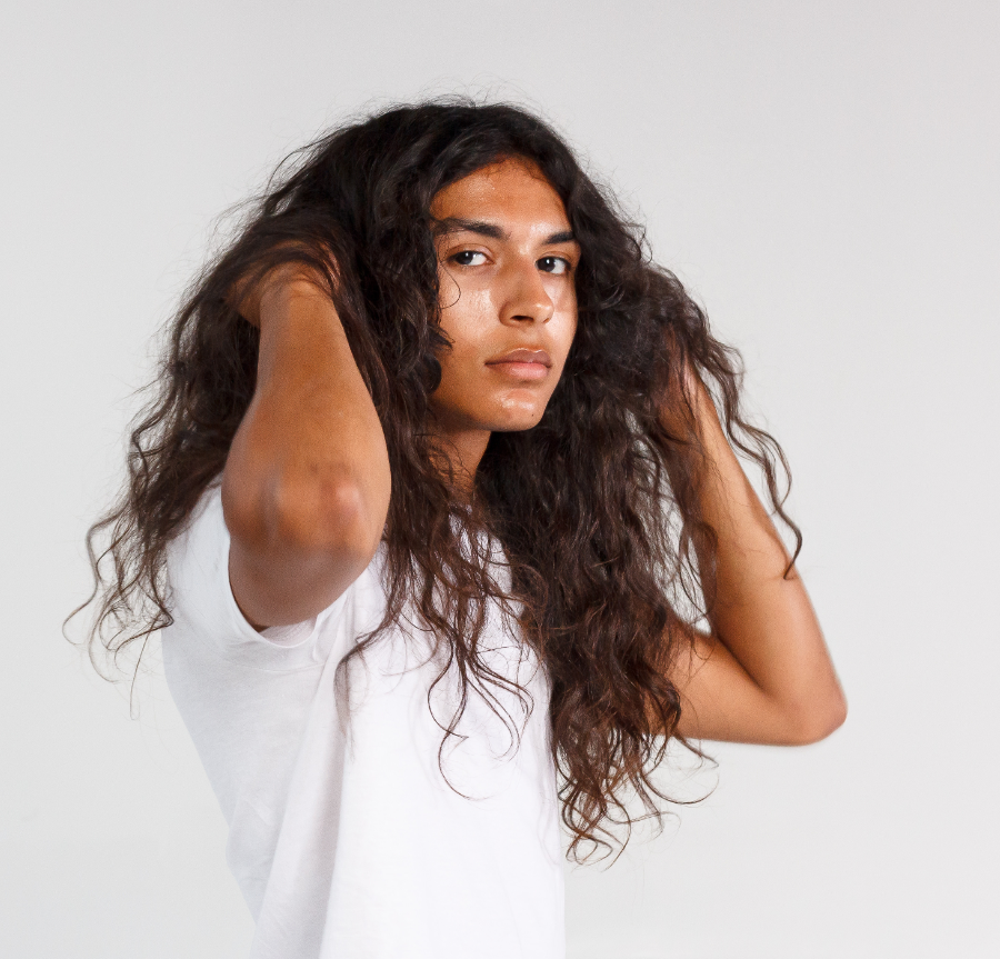 A man with long curly hair wearing a white t-shirt, looking at the camera with a neutral expression, set against a plain white background.