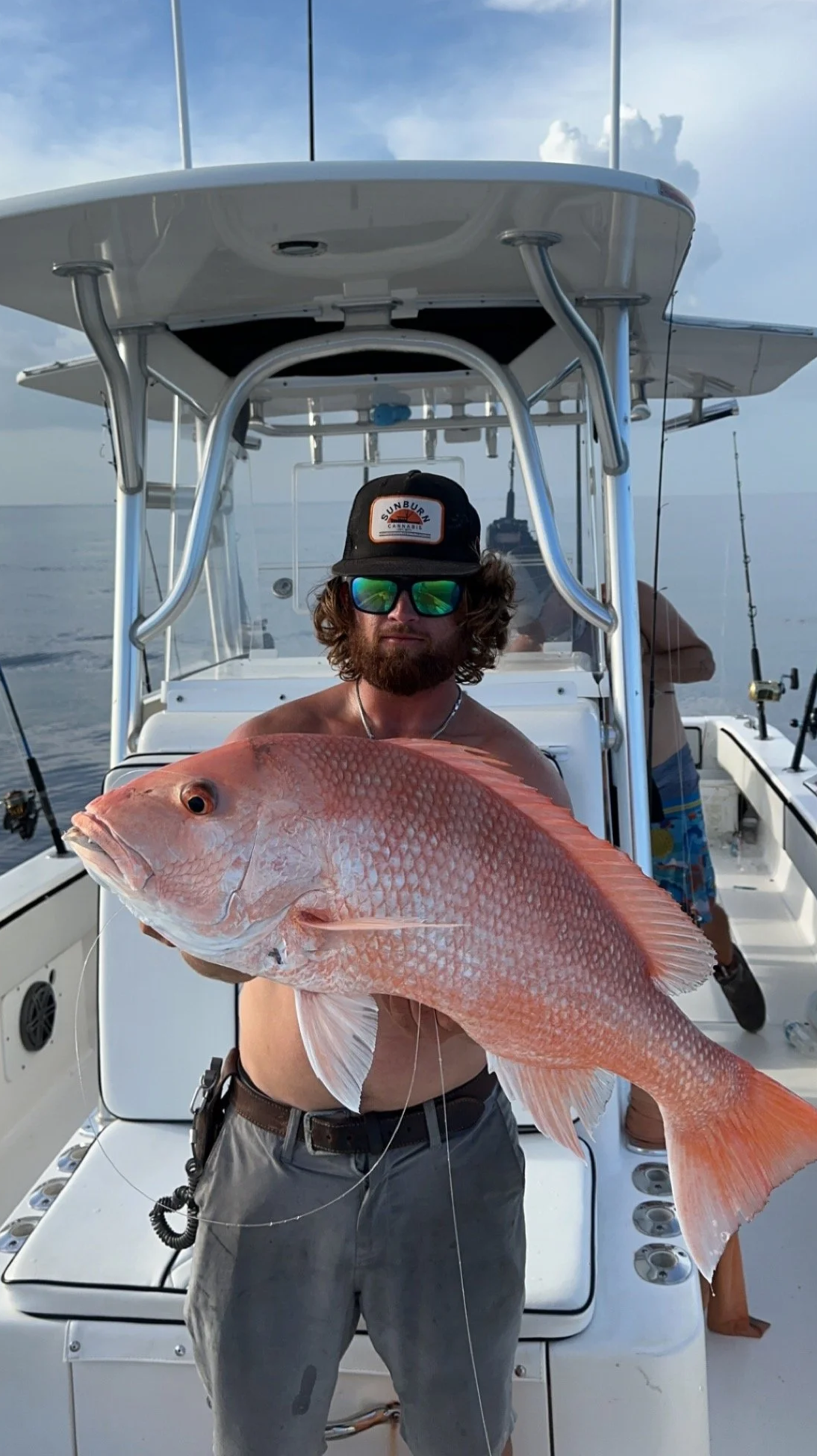 A man wearing sunglasses and a cap holding a large red fish on a boat during the day with calm water and cloudy sky in the background.