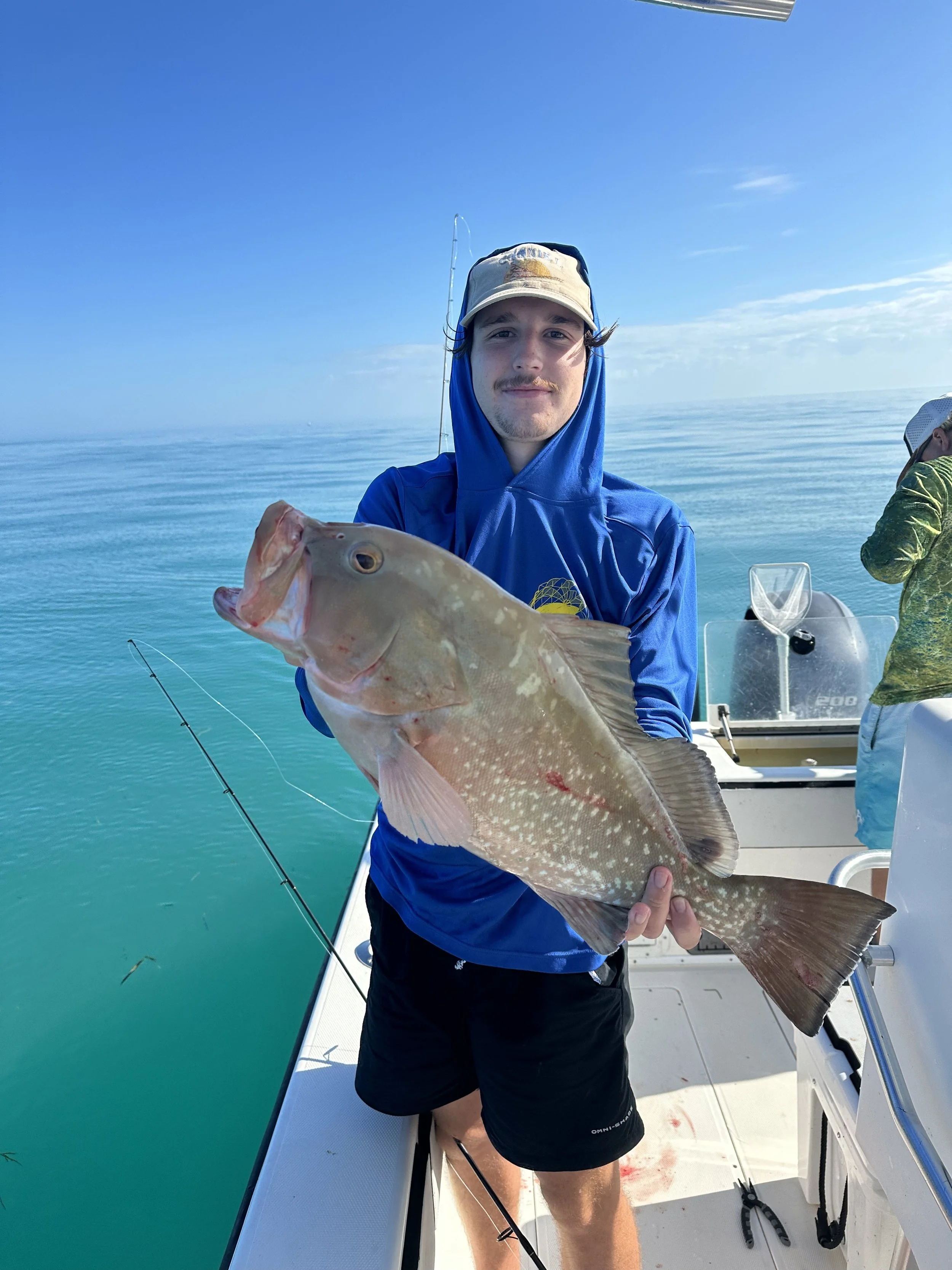 A man on a boat holding a large fish, with another person in the background, in a harbor or open water setting.