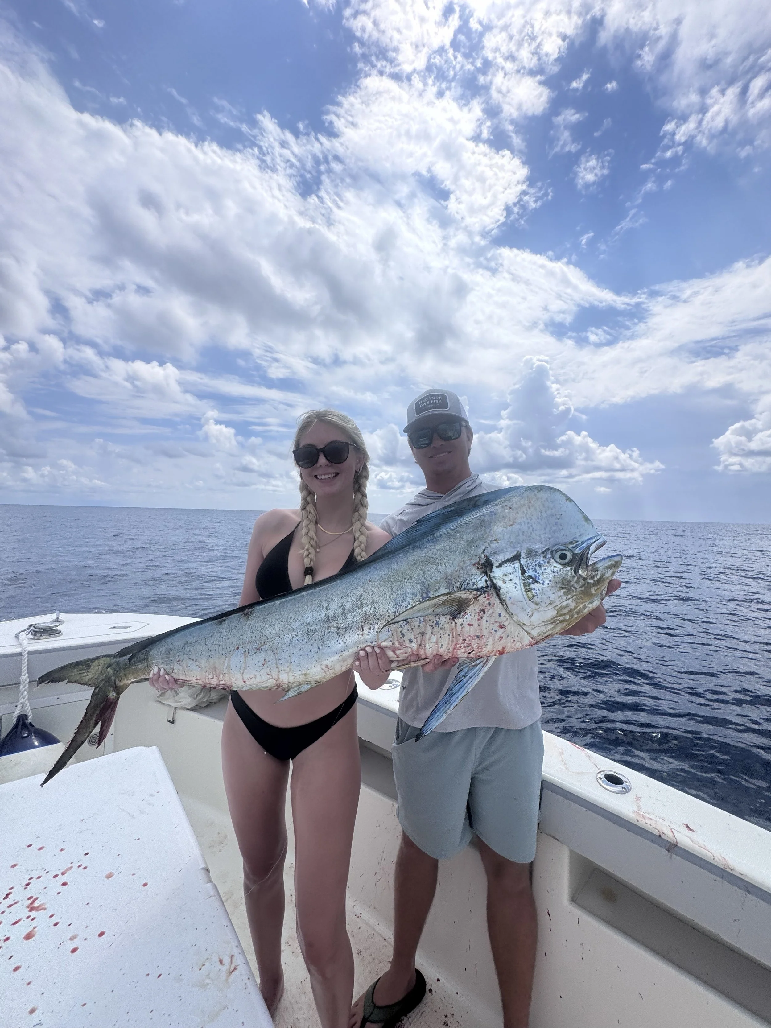 A woman and a man on a fishing boat holding a large fish they caught, with ocean and sky in the background.