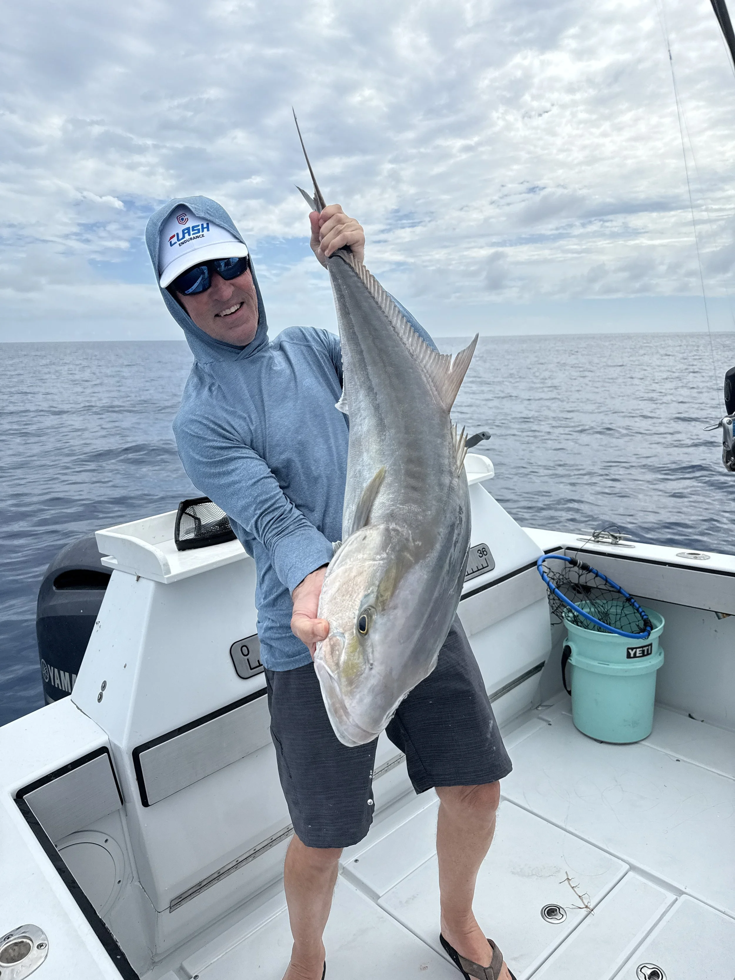 A man on a boat holding a large fish he caught, wearing a hoodie, sunglasses, and a baseball cap with a logo, with a sky and ocean background.