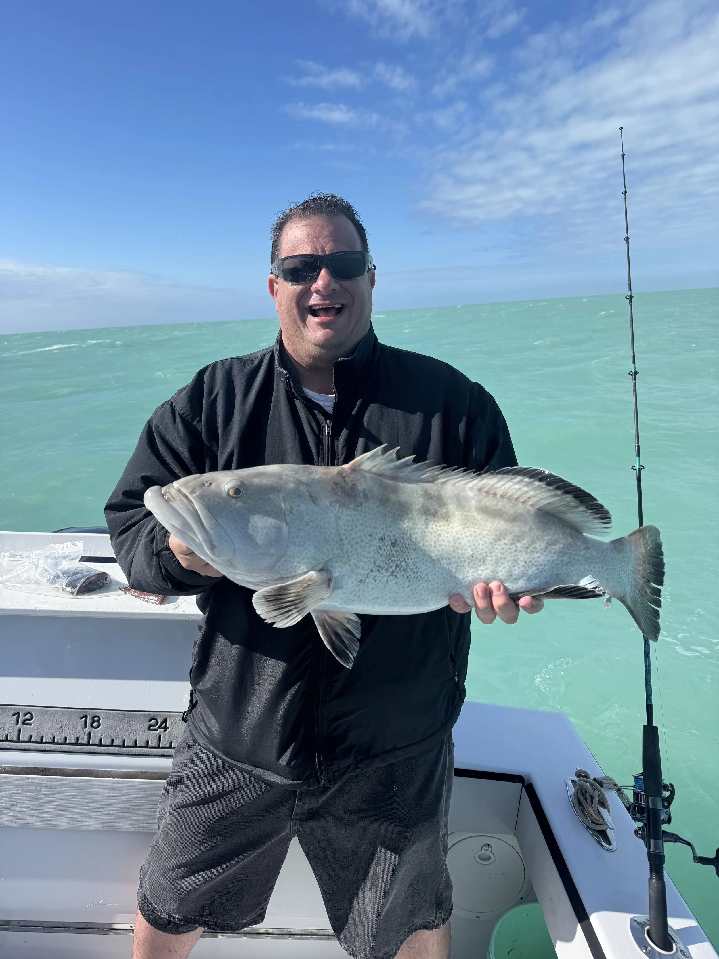 Man holding a large fish on a boat during daytime with ocean and sky in the background.