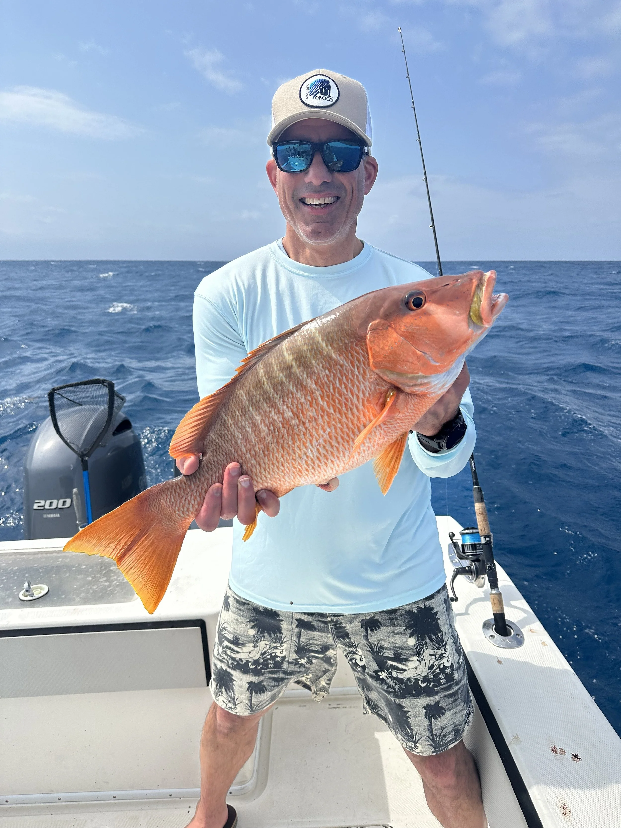 A man holding a large red fish on a boat in the ocean, smiling, wearing sunglasses, a baseball cap, a light blue shirt, and shorts with a tropical pattern.