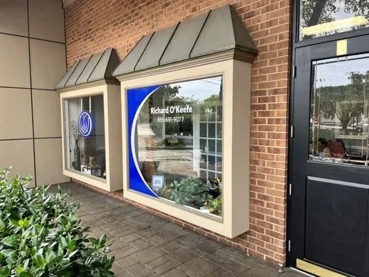 Storefront windows with a blue and white logo, one with the name 'Richard O'Keefe' and contact number, beside a black door, with bushes in front.