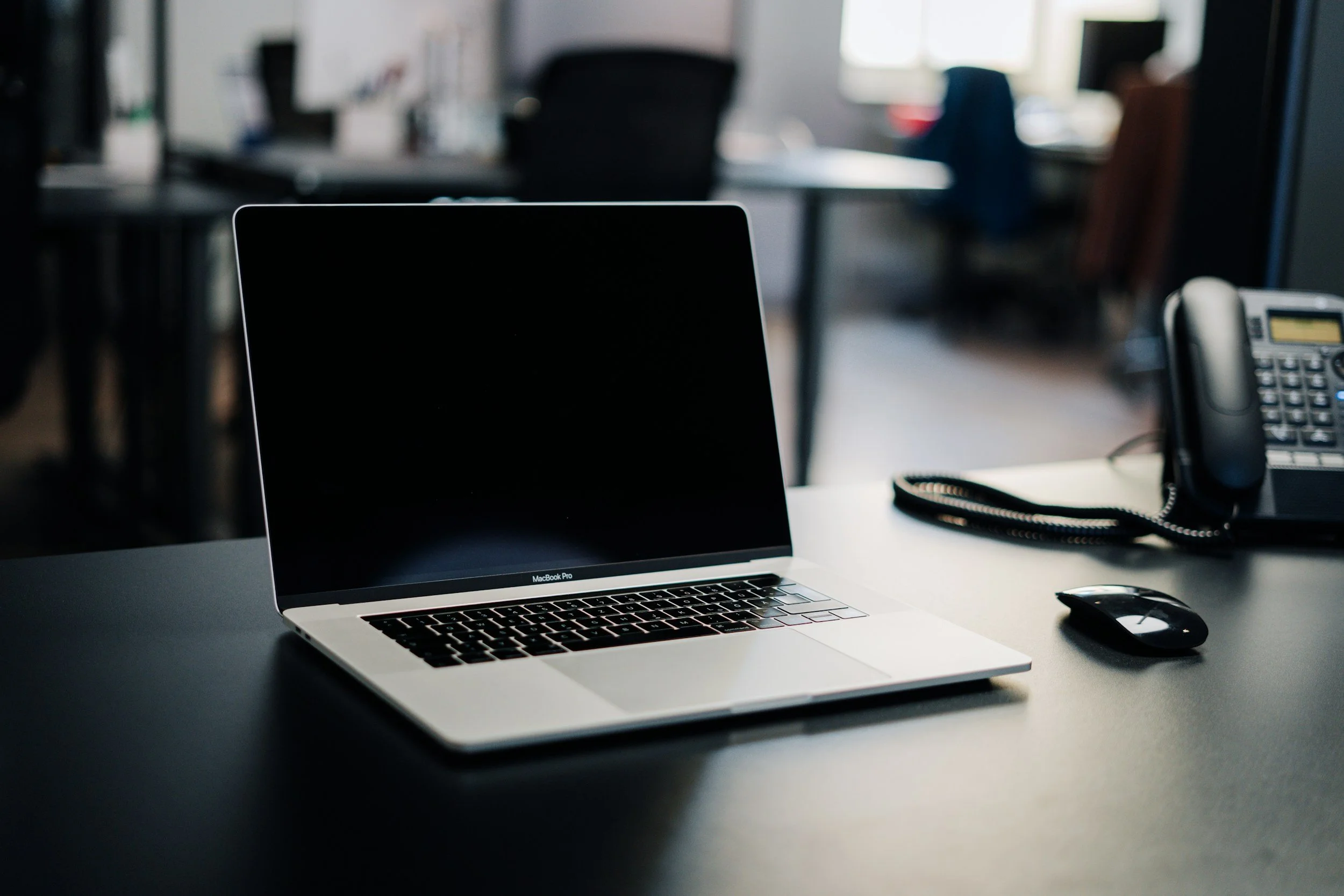A workspace with a silver MacBook Pro on a white desk, a black desk phone, and an office environment with desks and chairs in the background.