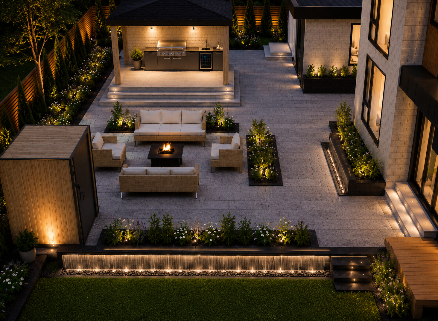 A modern outdoor patio at night with a seating area, fire pit, and built-in kitchen, illuminated by landscape lighting and surrounded by lush greenery.