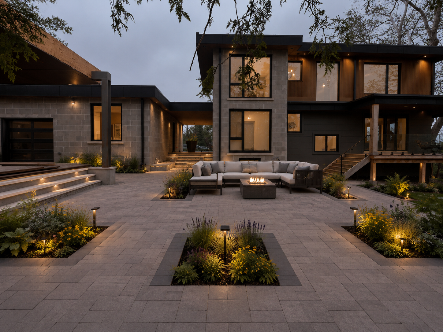 Modern house exterior at dusk with a paved patio, outdoor furniture, fire pit, illuminated garden beds, and large windows.