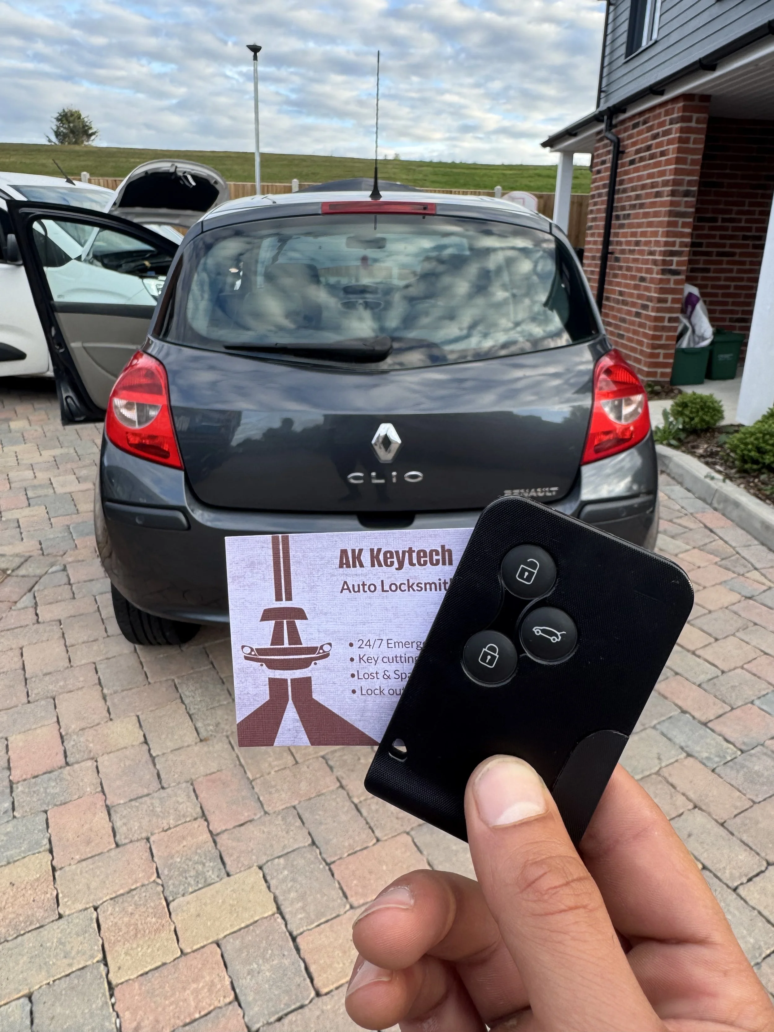 A person holding a car key fob with a black plastic casing and four buttons, in front of a black Renault Clio parked outside on a paved driveway. The car's rear window reflects the cloudy sky, and there's a sign on the ground with the text 'AK Keytec