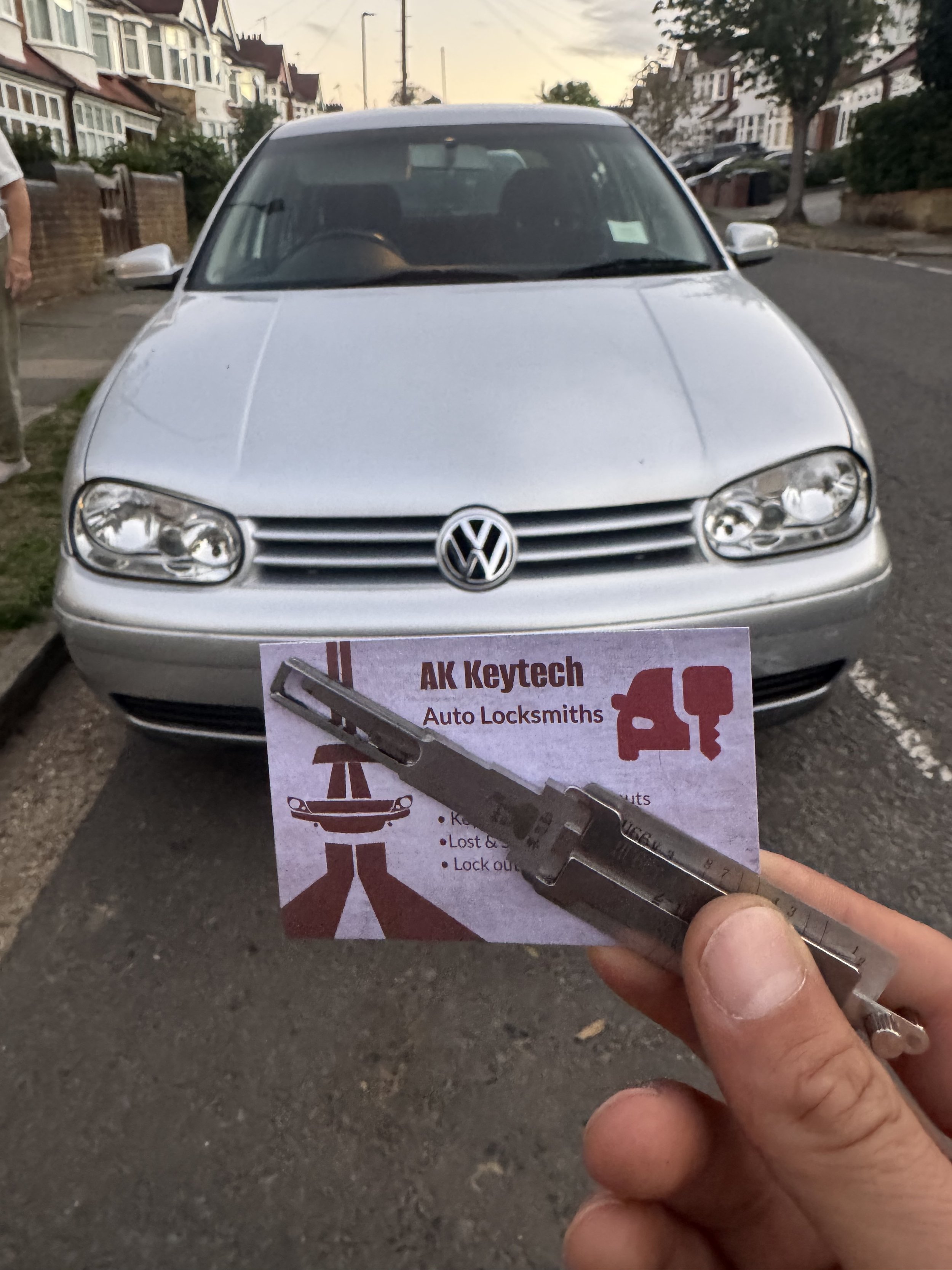 Front view of a silver Volkswagen car parked on a residential street during early evening, with a person holding a key and a flyer for a locksmith business.