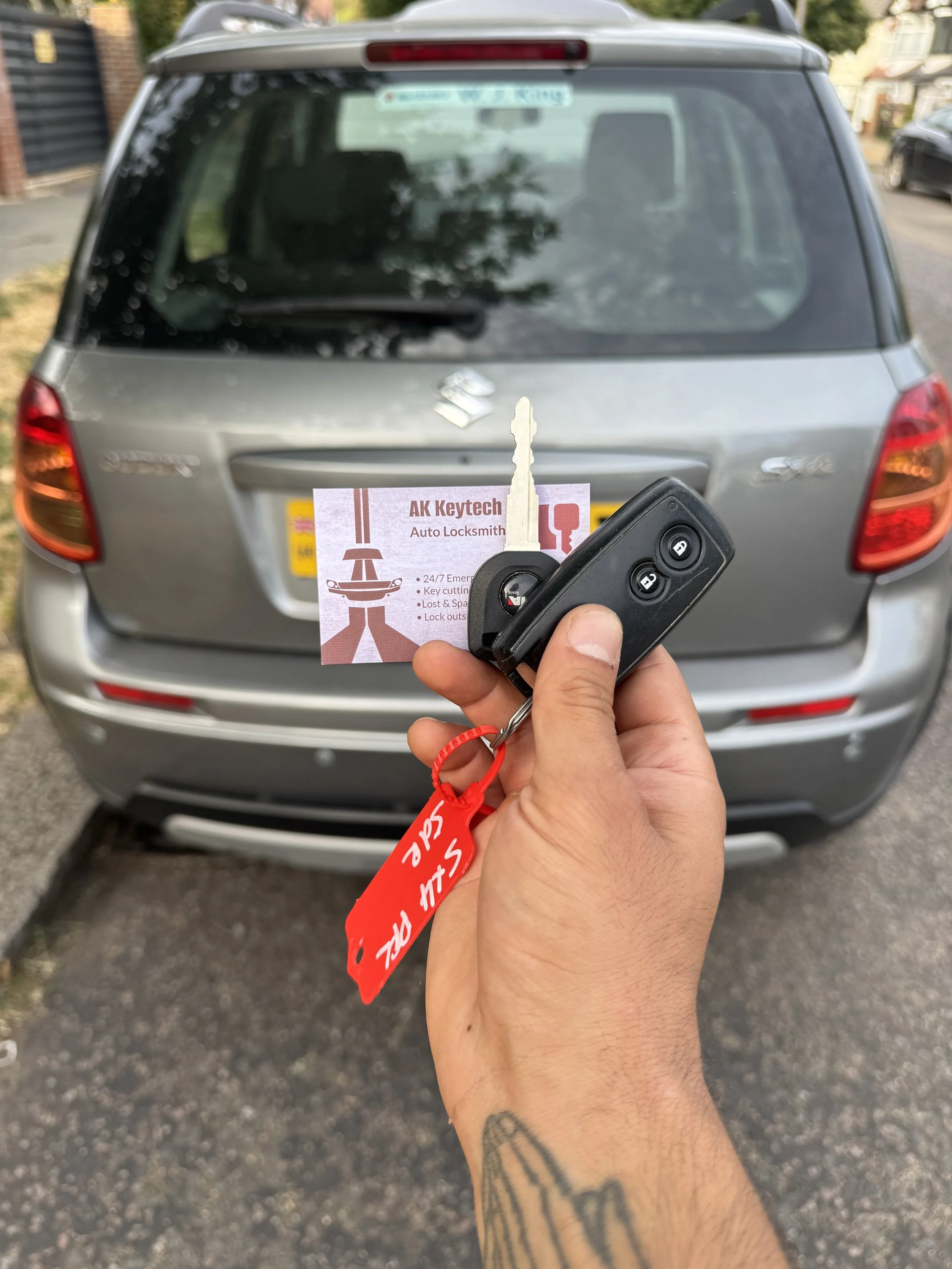 Hand holding a car key and a business card for AK Keytech Auto Locksmith in front of a silver SUV.