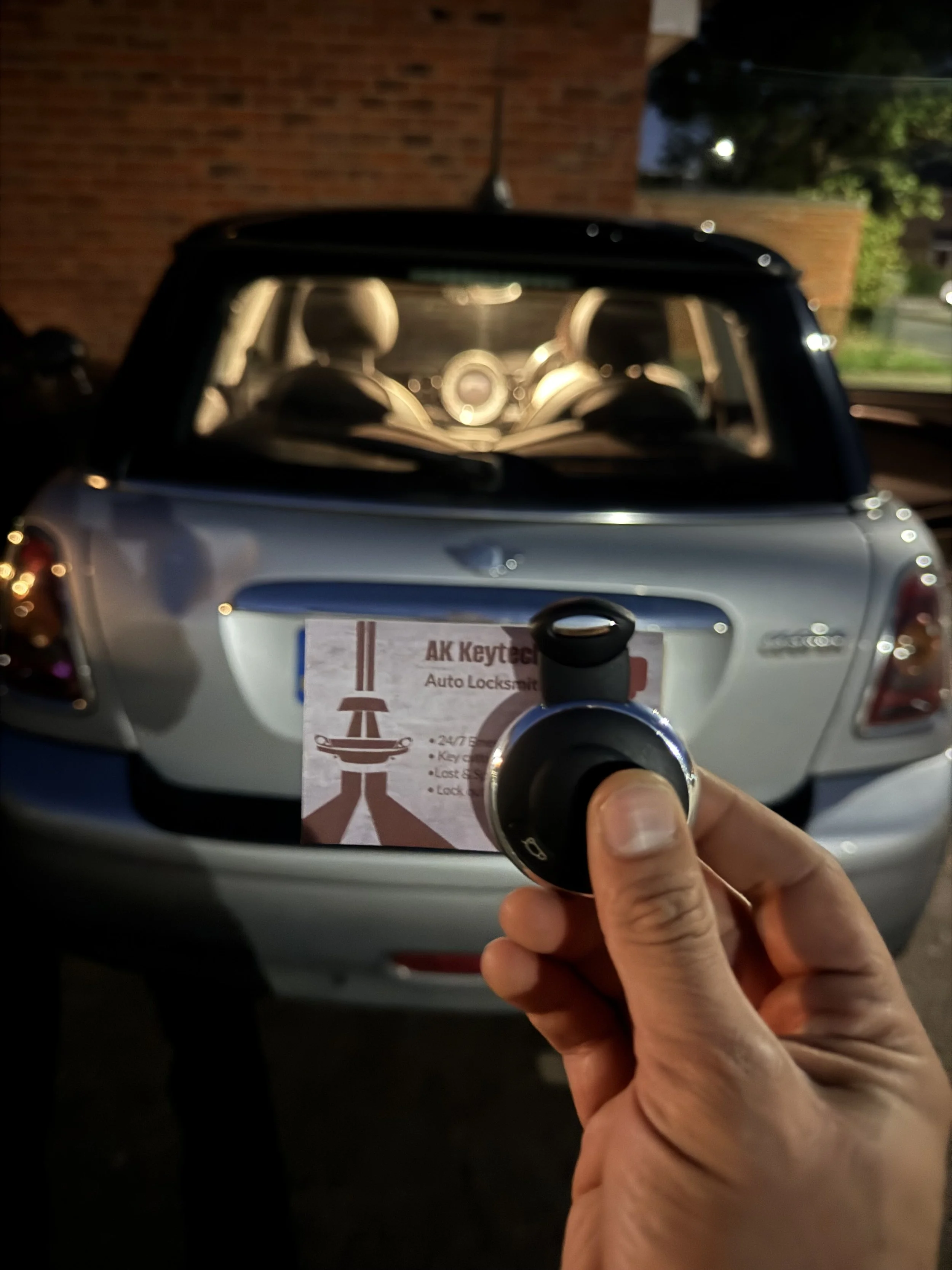 Person holding a car key fob in front of a rearview mirror reflecting the back of a silver car with a business card or flyer for "AK Keytech Auto Locksmith".