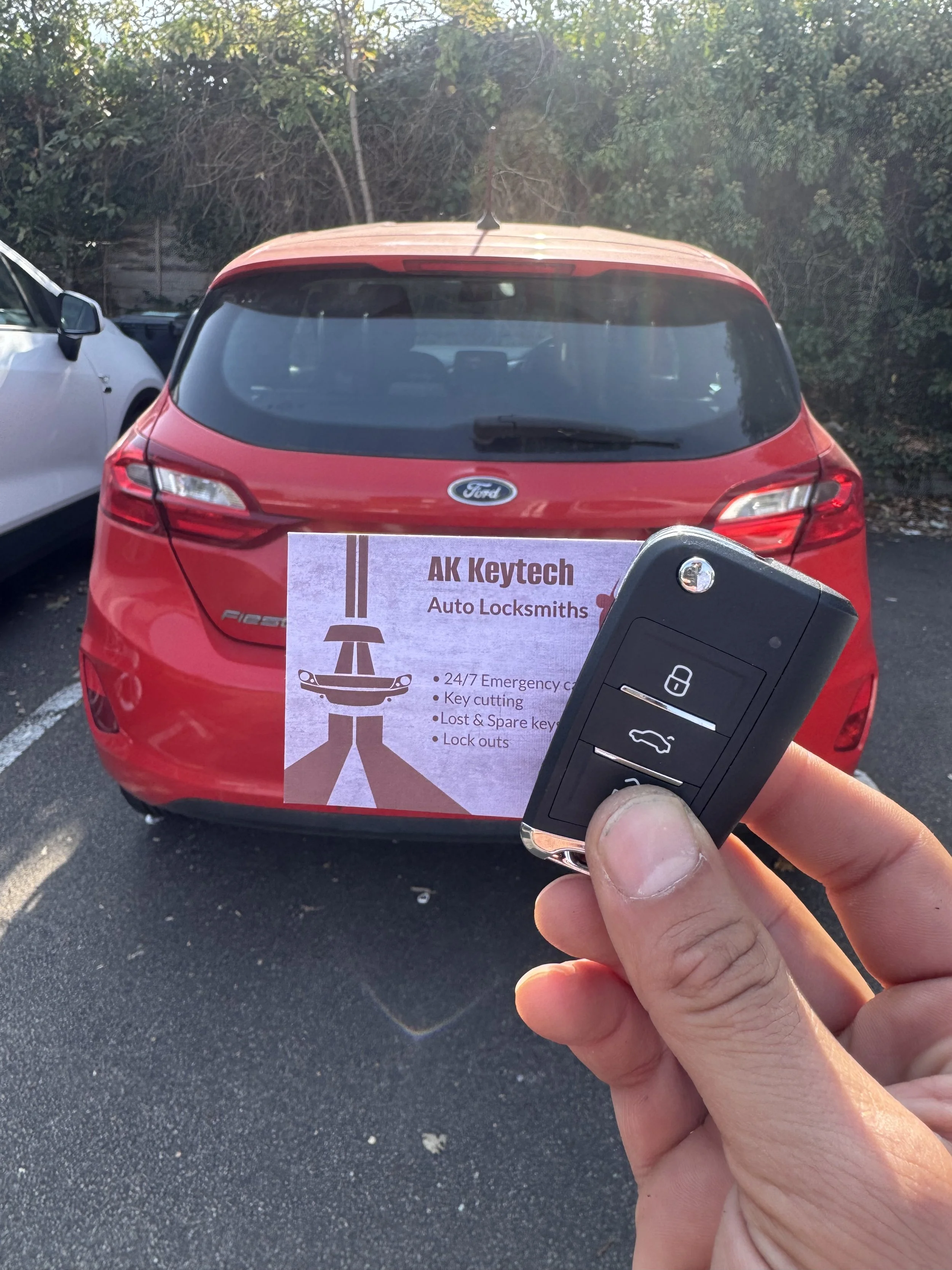 Red hatchback car parked outdoors with a vending machine advertisement in front, showing a key fob remote control in a person's hand.