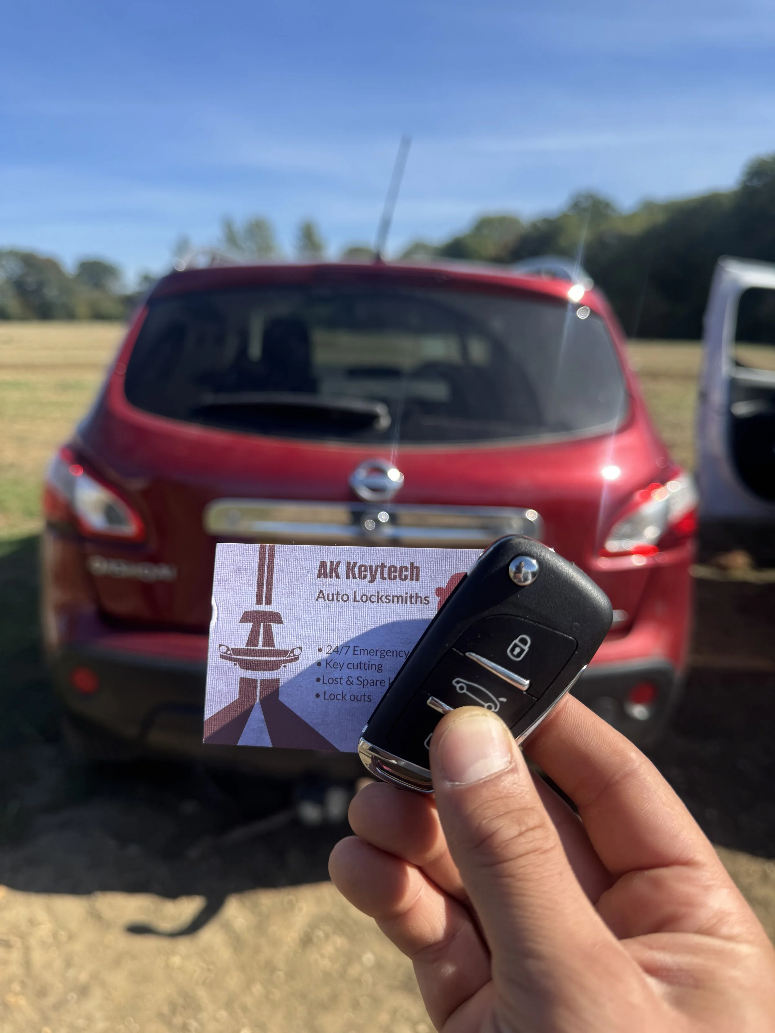 Person holding a car key fob with a business card for AK Keytech Auto Locksmiths, standing in front of a red Nissan SUV parked outdoors on a sunny day.