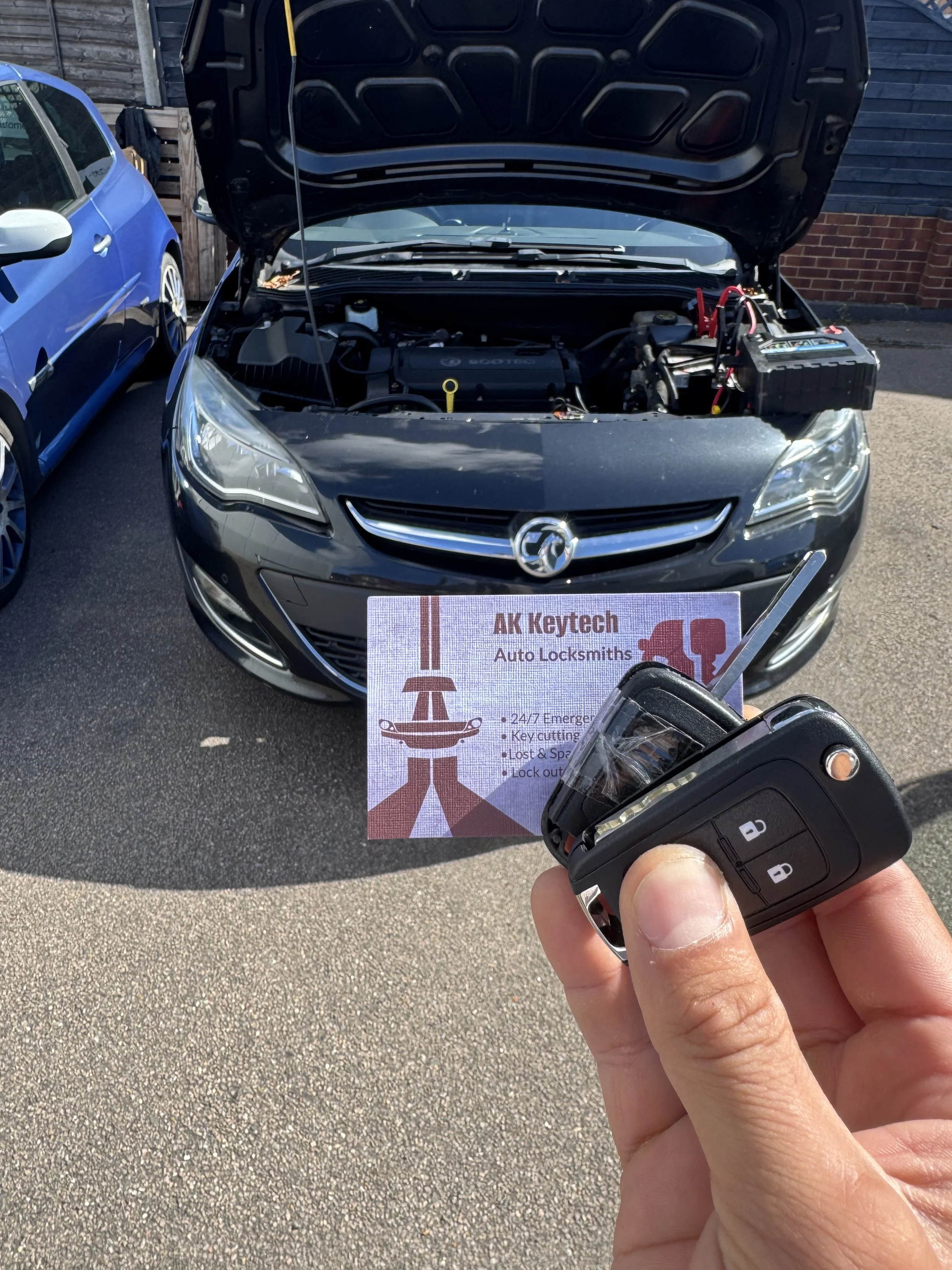 A person holding a car key fob in front of a black car with its hood open, parked in a lot next to a blue car. There is a sign on the front of the black car that reads 'AK Keytech Auto Locksmiths' with additional services listed below.