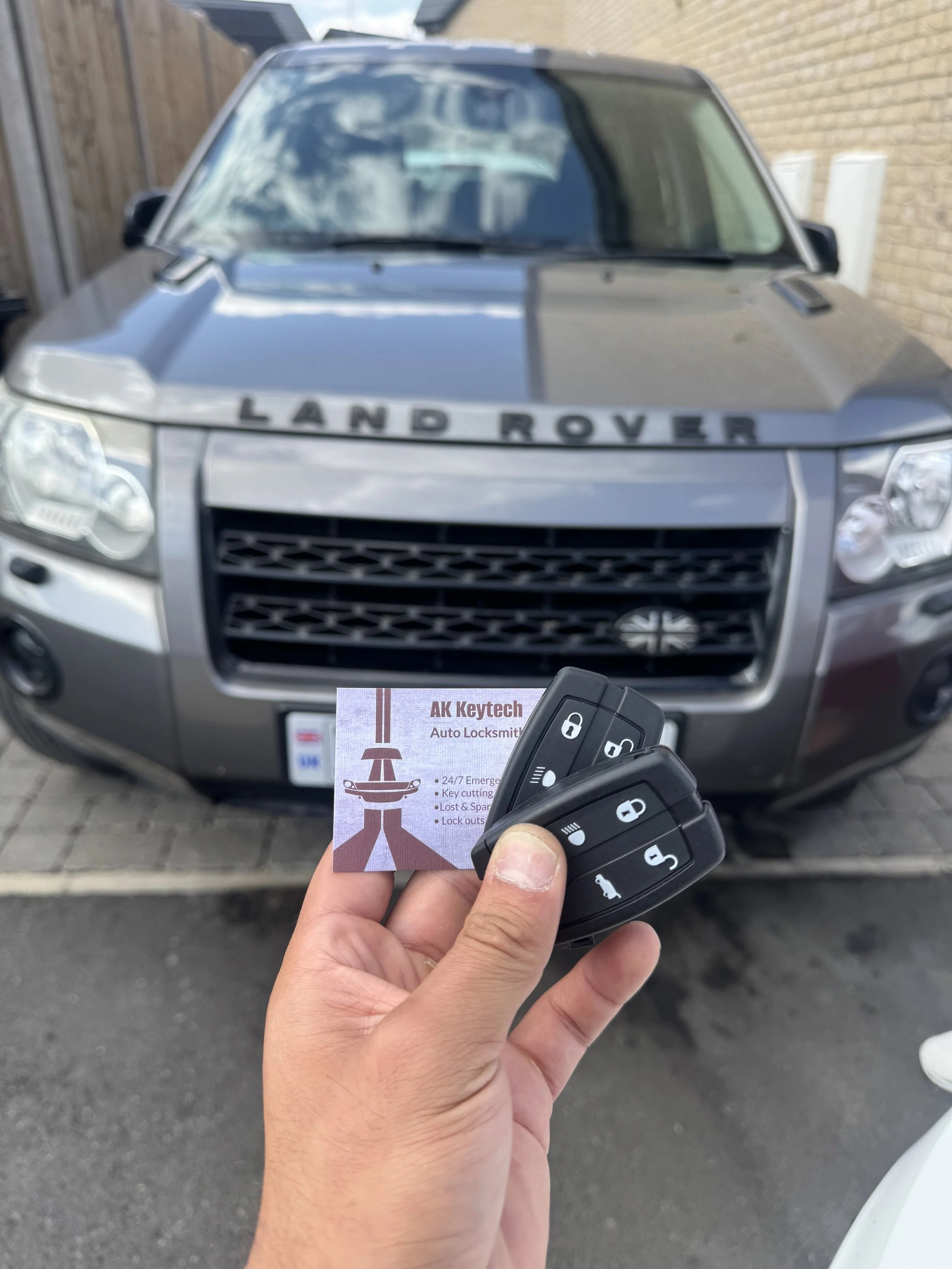 Person holding car key fob and business card in front of a gray Land Rover vehicle parked outside with brick wall and wooden fence in the background.