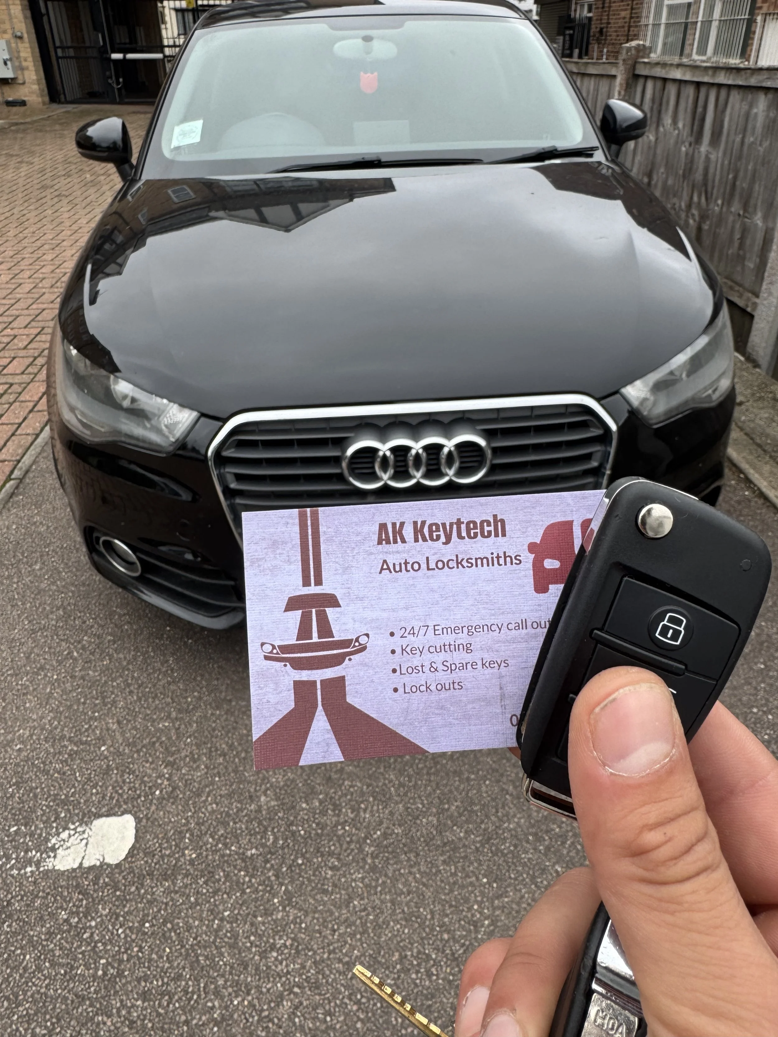 Front view of a black Audi car parked on a driveway with a sign for AK Keytech Auto Locksmiths held up in front. The sign mentions services like 24/7 emergency calls, key cutting, lost and spare keys, and lockouts. A car key remote is held in the han