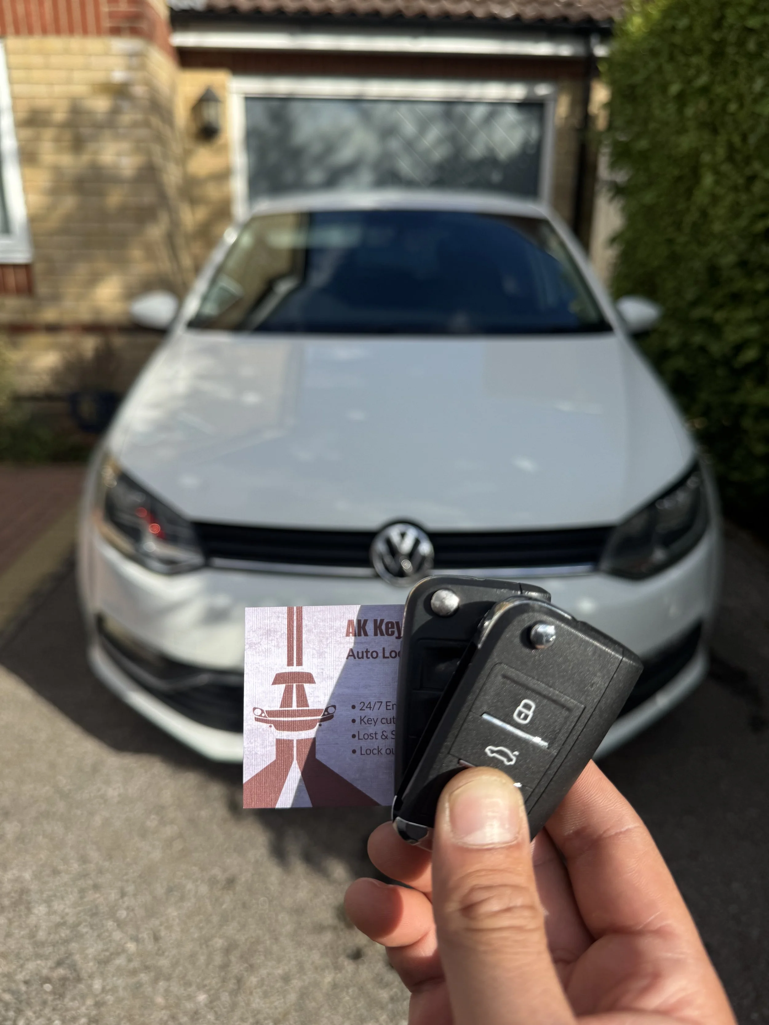 A person holding a car key fob and a key card in front of a white Volkswagen parked outside a house.