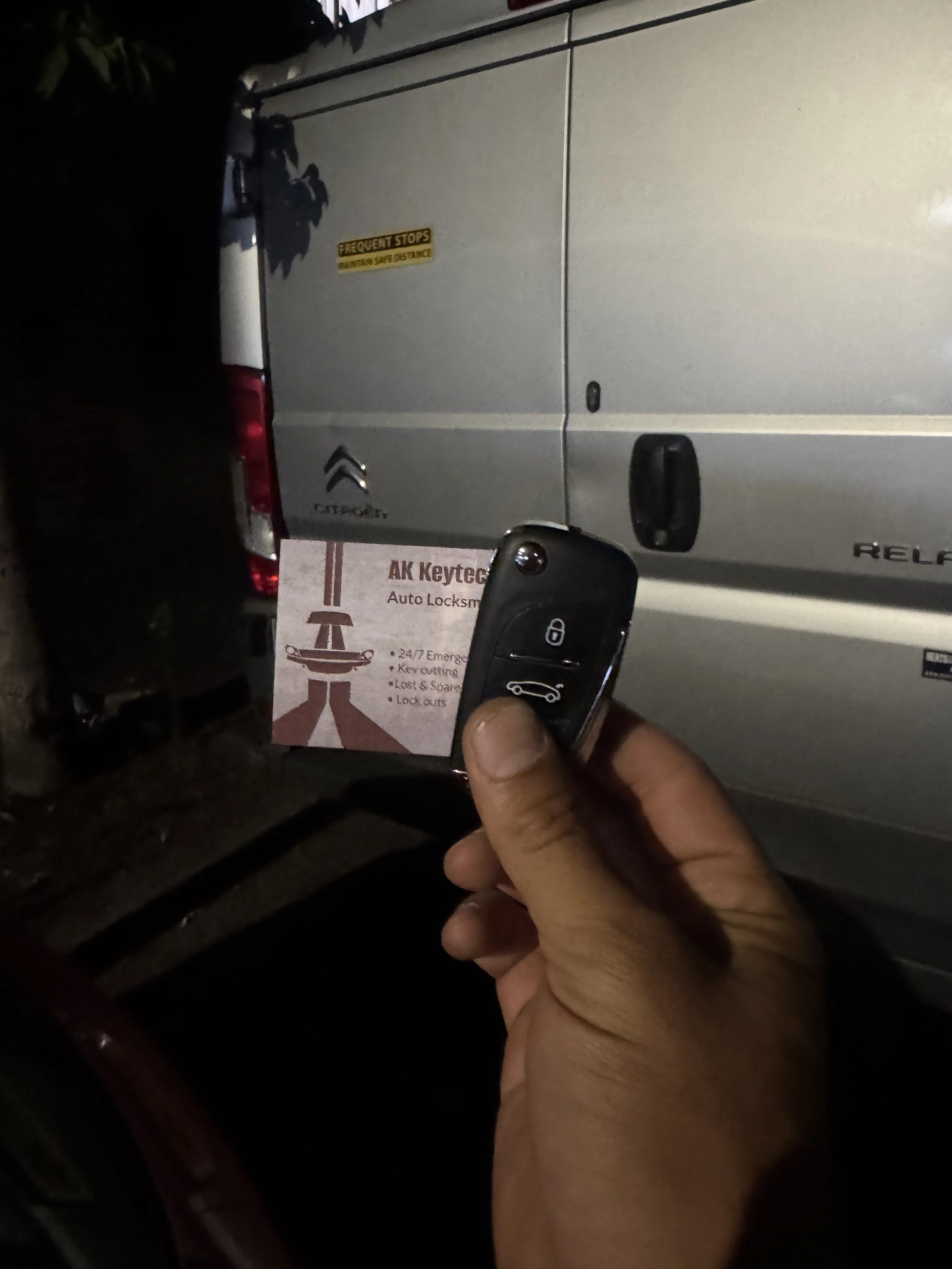 A person holding a car key in front of a silver van at night. The van has a yellow warning sticker that reads "FREQUENTLY STORES" and a logo of a Citroën vehicle. There is a sign in front of the van that reads "AK Keytec Auto Locksmith" with addition