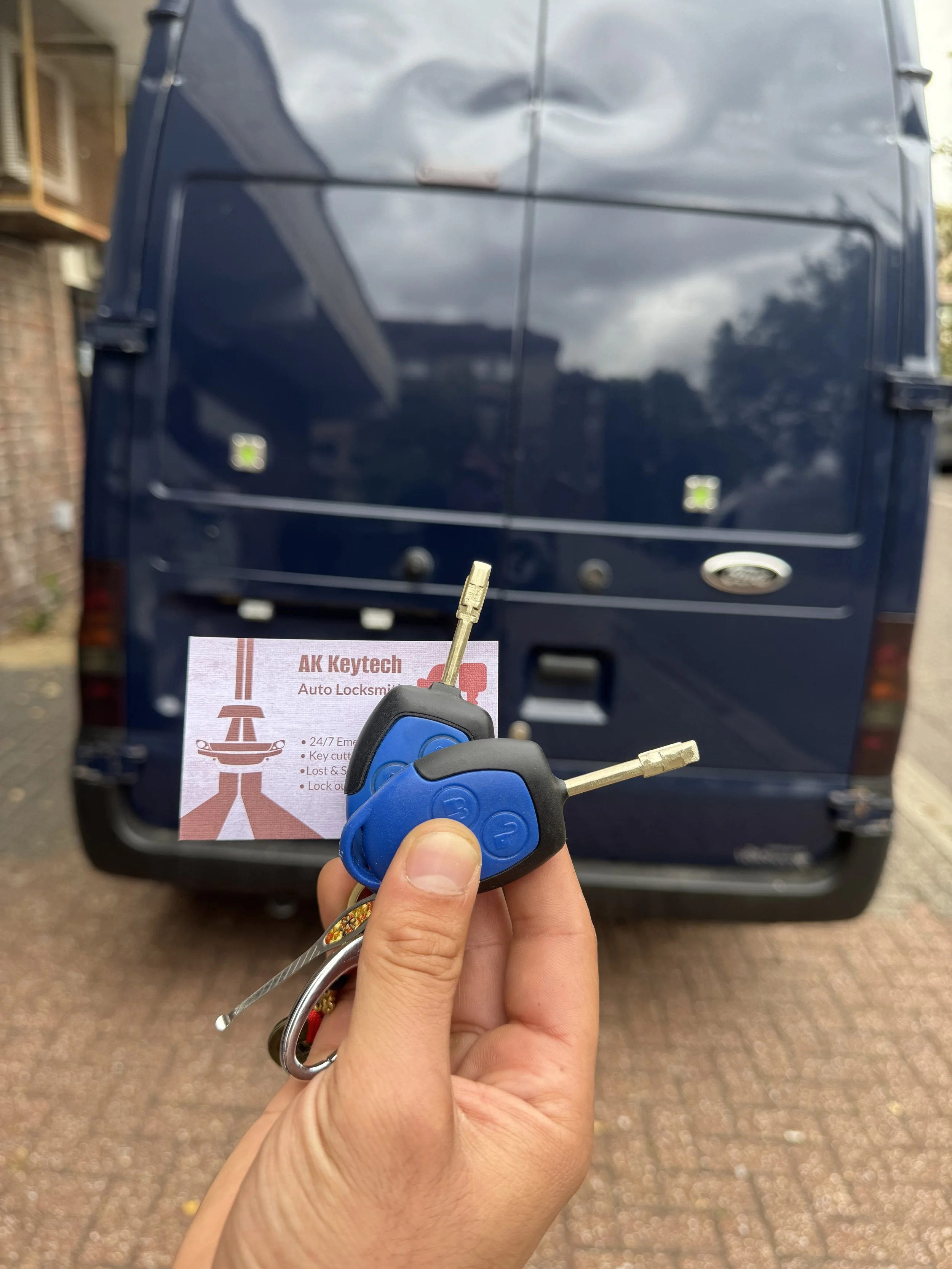 Person holding a blue car key fob with a key, in front of a dark blue vehicle, with a business card for AK Keytech auto locksmith in the background.