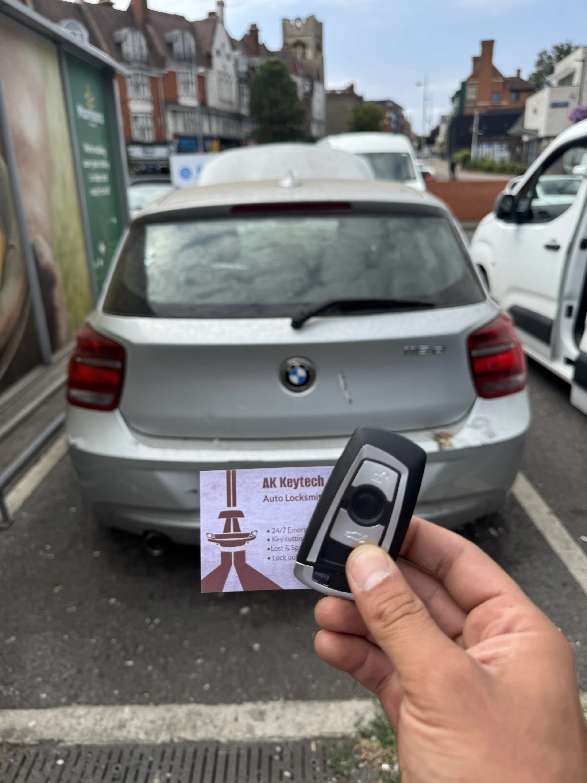 Person holding a car remote in front of a silver BMW parked in a parking lot with other vehicles and buildings in the background.