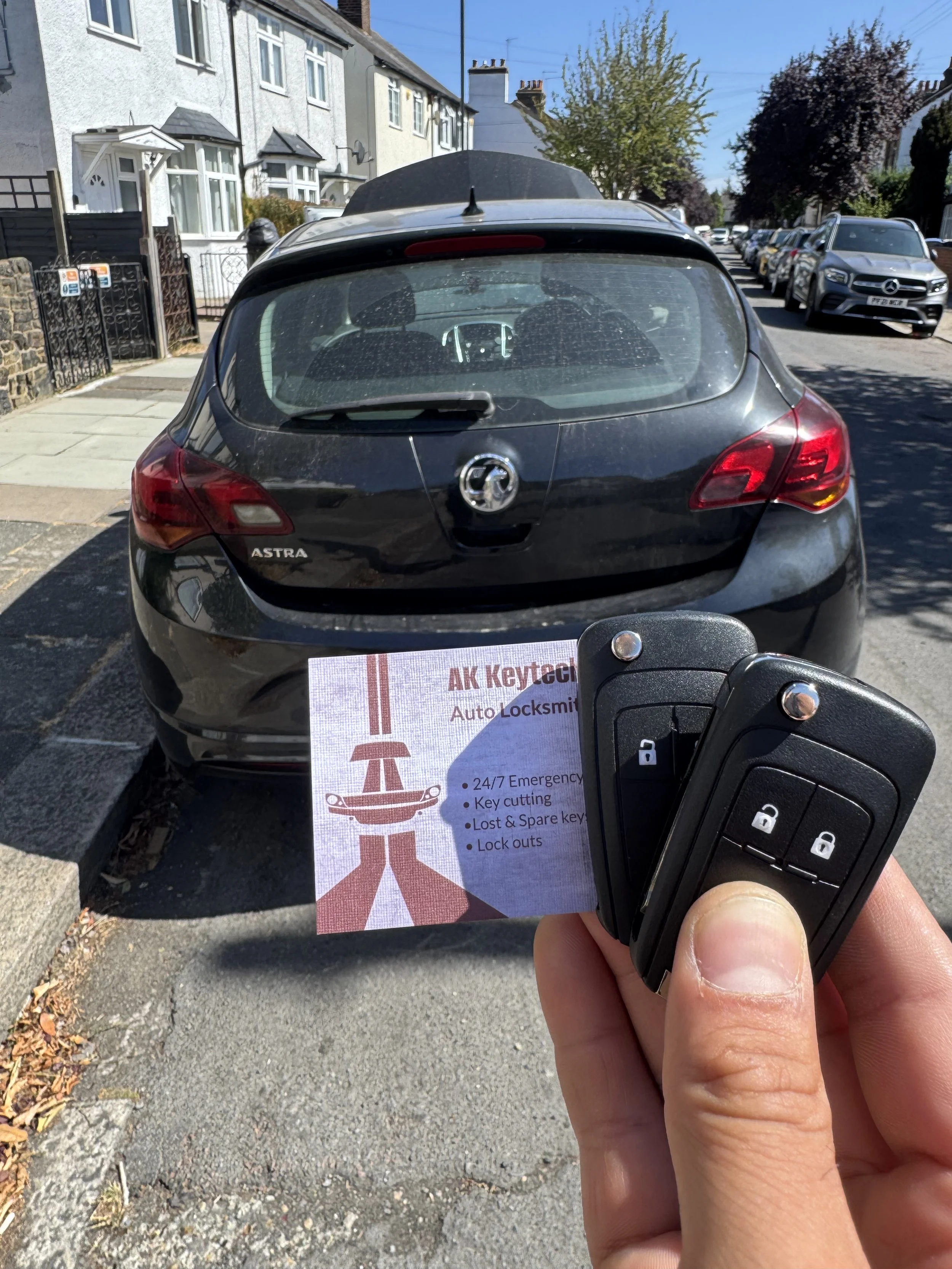 Person holding a car key fob in front of a black Vauxhall Astra parked on a residential street with multiple parked cars and houses.