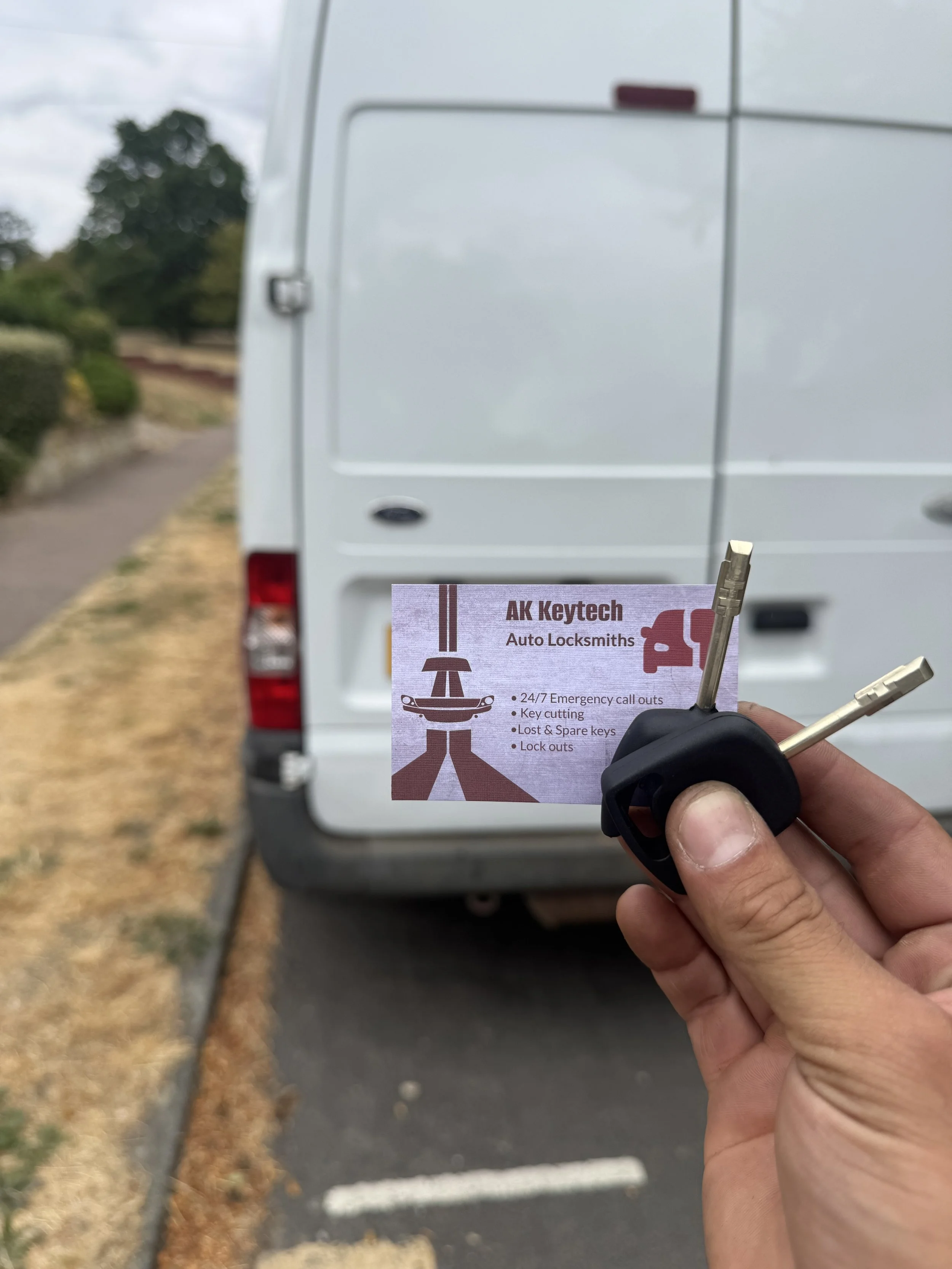 Hand holding a car key in front of a white van, with a business card for AK Keytech Auto Locksmiths visible.