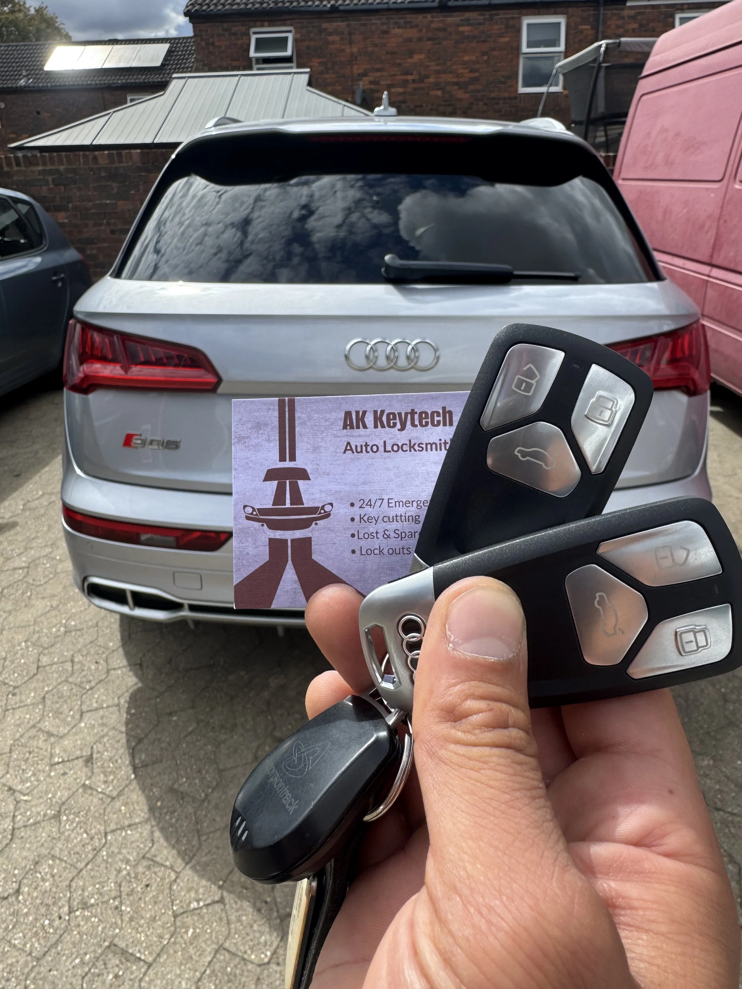 Person holding a car key fob in front of a silver Audi SUV with a sign for AK Keytech Auto Locksmith in the background.